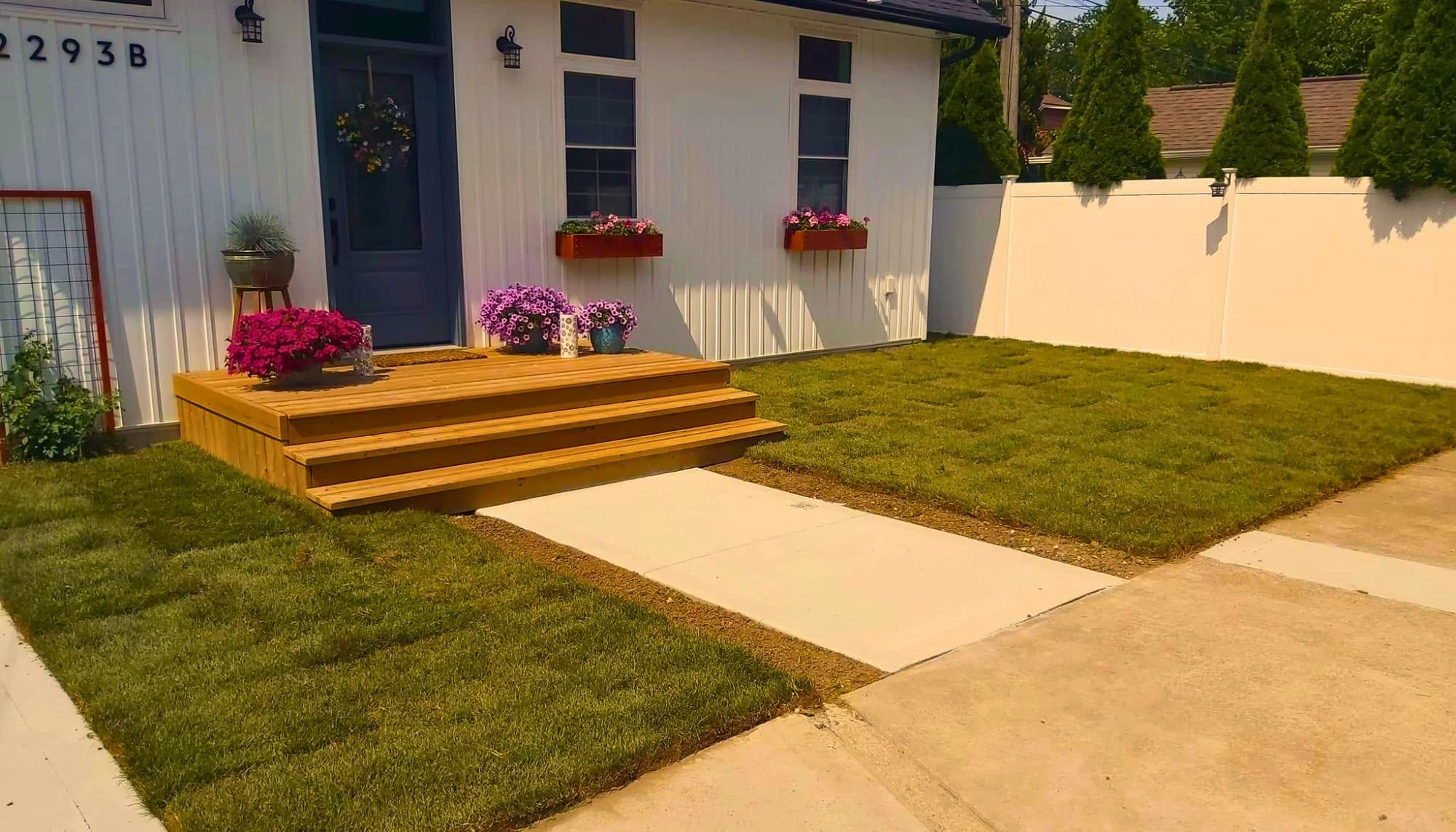 A small front yard with a wooden staircase leading to a blue door on a white house, decorated with potted flowers and hanging baskets, surrounded by a green lawn, concrete pathway, and a white fence with trees in the background.