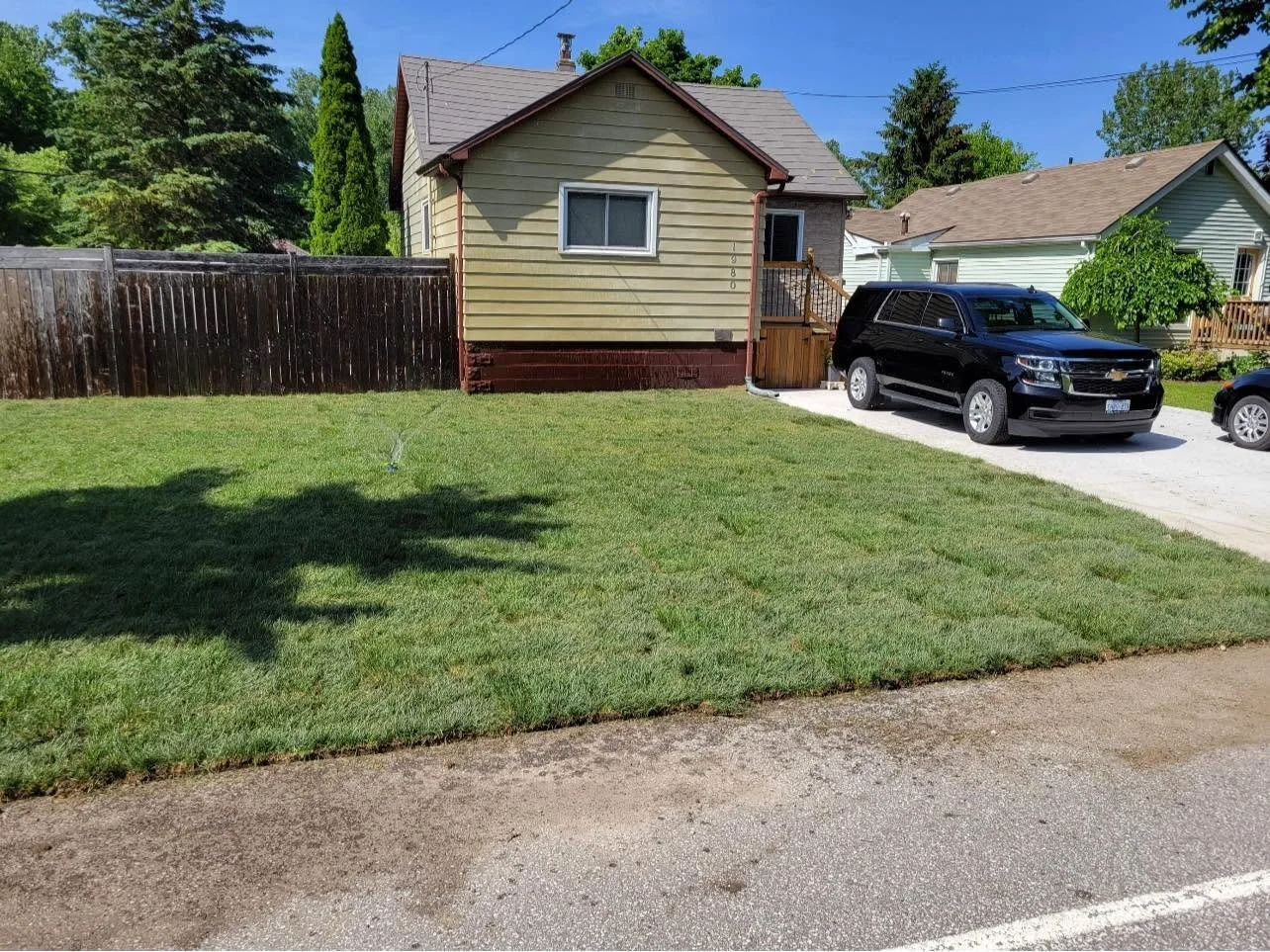 A backyard with a small house, a wooden fence, a well-maintained lawn, and a parked black SUV on a concrete driveway.