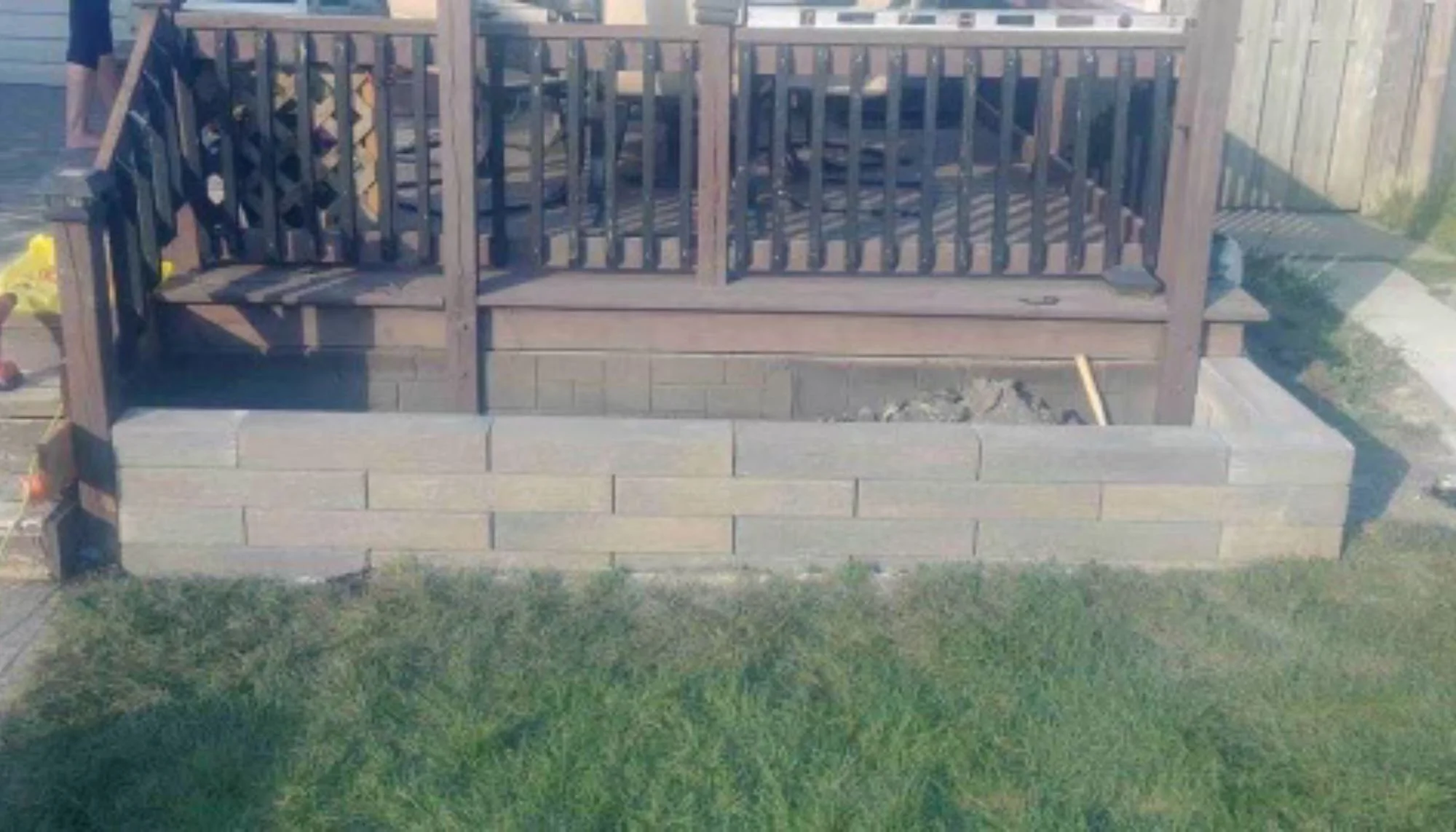 Image of a wooden deck with a railing, elevated above a stone foundation, with grass in the foreground.
