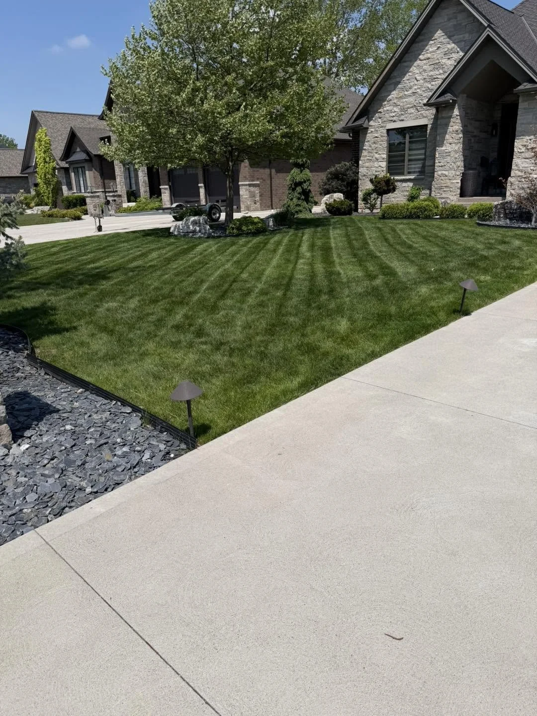 A well-maintained front yard with a lush green lawn, a tree, decorative rocks, and landscaped bushes in front of a modern house with stone and brick exterior, under a clear blue sky.