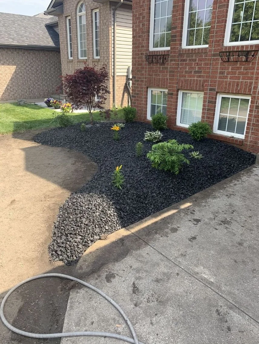 Clean landscaped garden in front of a brick house with small plants, bushes, and a prayer garden, bordered with black mulch, and part of a wooden sidewalk.
