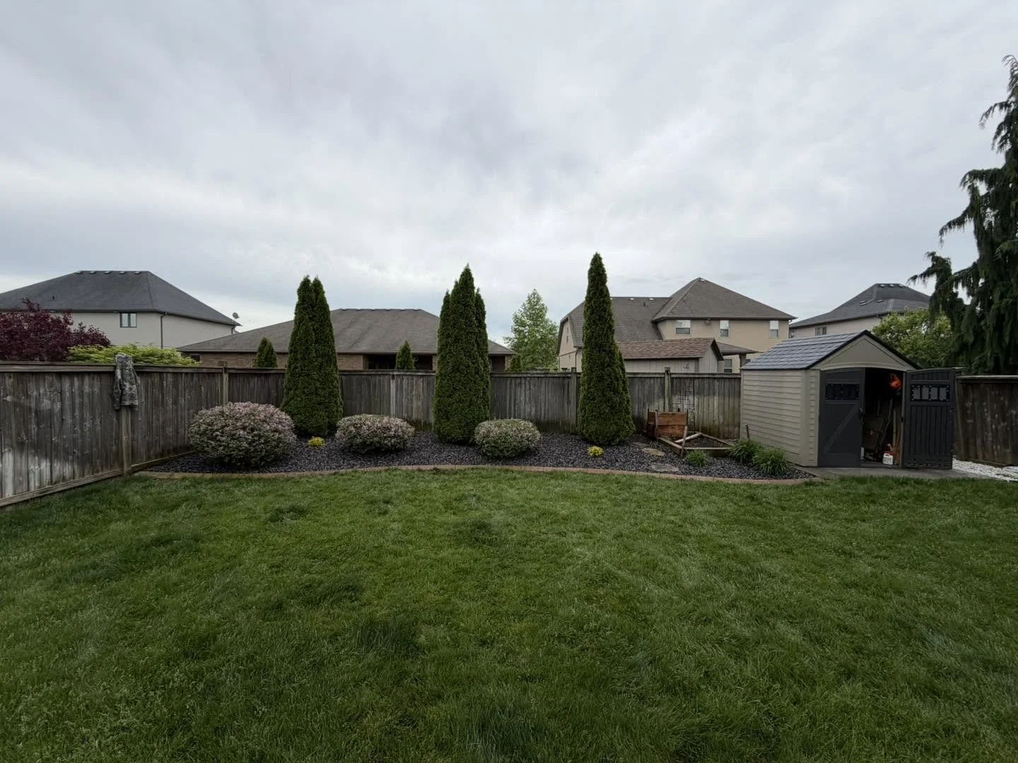 Backyard with manicured lawn, wooden fence, shrubbery, tall shaped trees, garden shed, and neighboring houses under a cloudy sky.