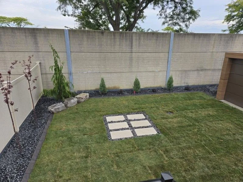 A small backyard with neatly trimmed grass, a border of black gravel, and a concrete fence. There are small plants and rocks along the fence, and four rectangular stepping stones in the grass. A part of a wooden shed or structure is visible on the right.