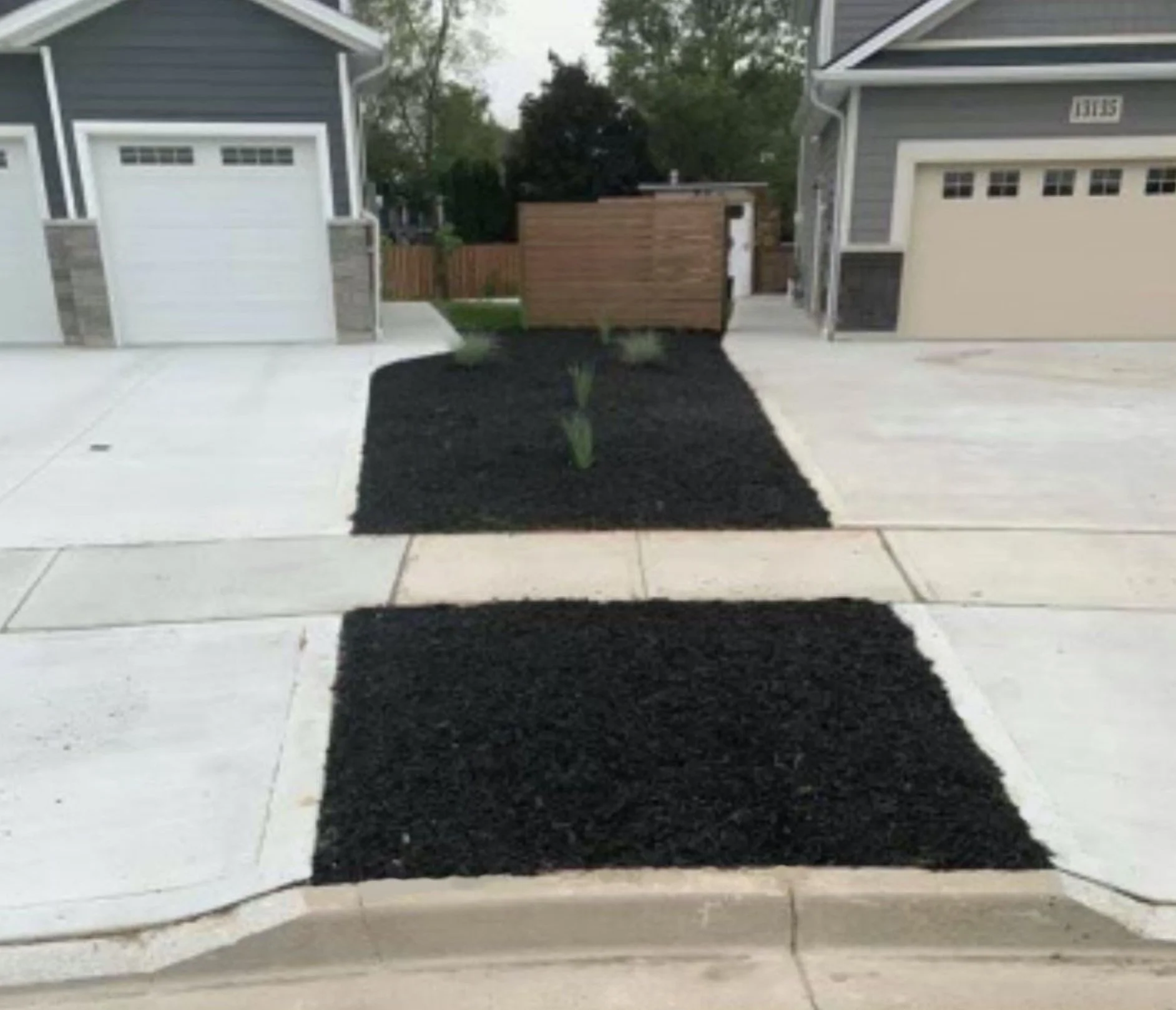 Front view of a residential driveway with a black mulch bed featuring young plants, flanked by two gray garage doors on either side.