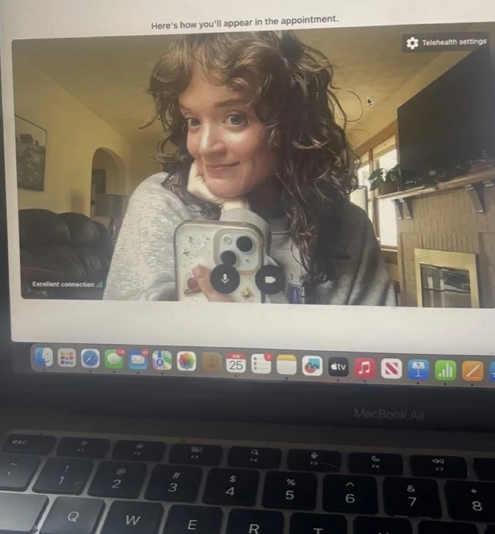 A woman taking a selfie during a video call on her MacBook Air, with a cozy living room in the background. She has curly hair and is smiling at the camera.
