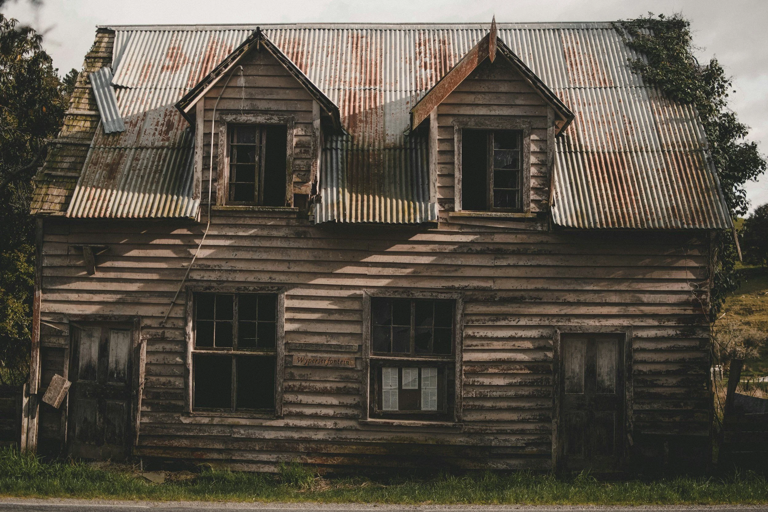 Old, weathered two-story wooden house with a rusted corrugated metal roof and multiple broken or missing windows. The house appears abandoned and dilapidated.