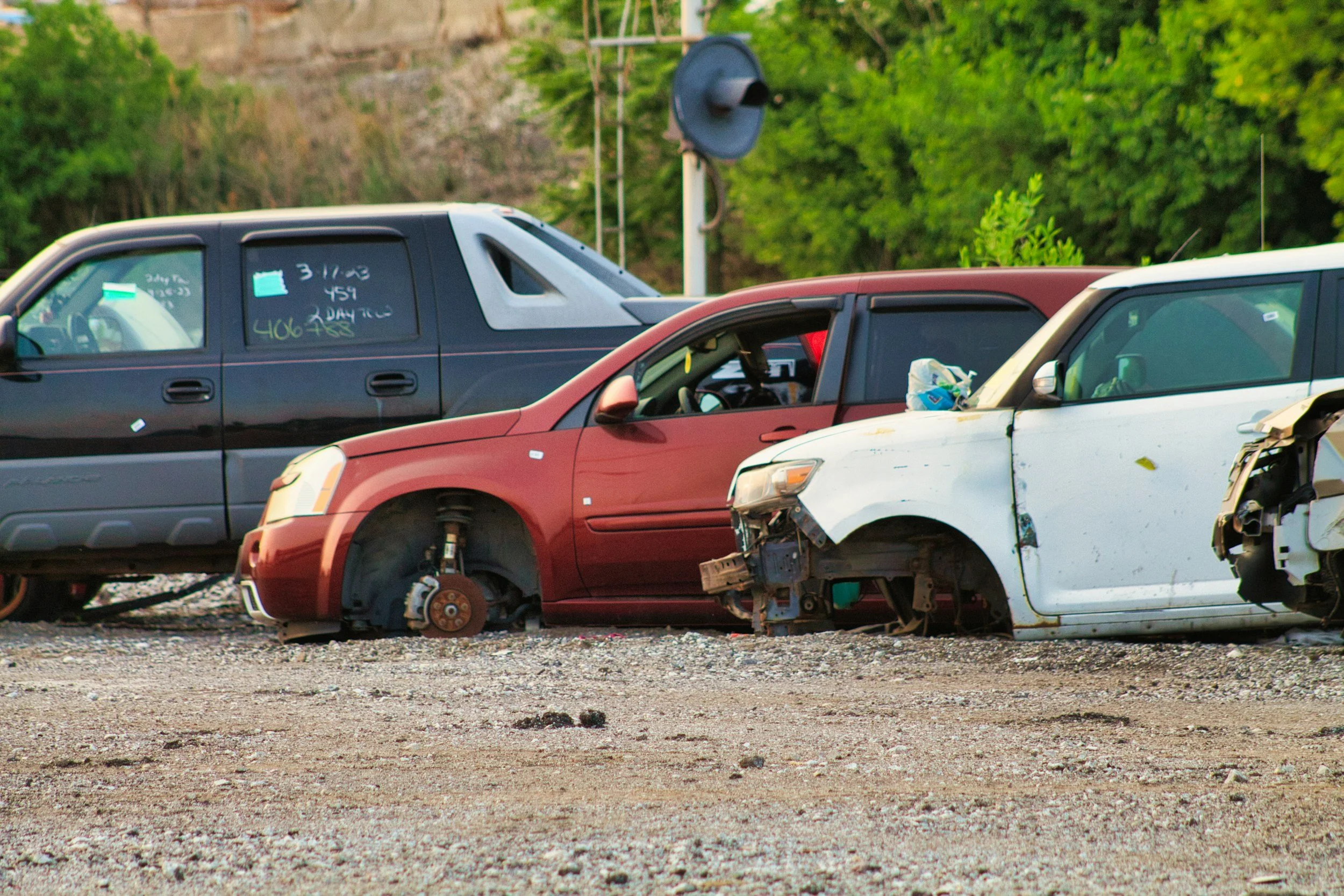 A row of damaged cars, with one missing wheels and another with front parts missing, sit on a gravel lot, likely in a junkyard.