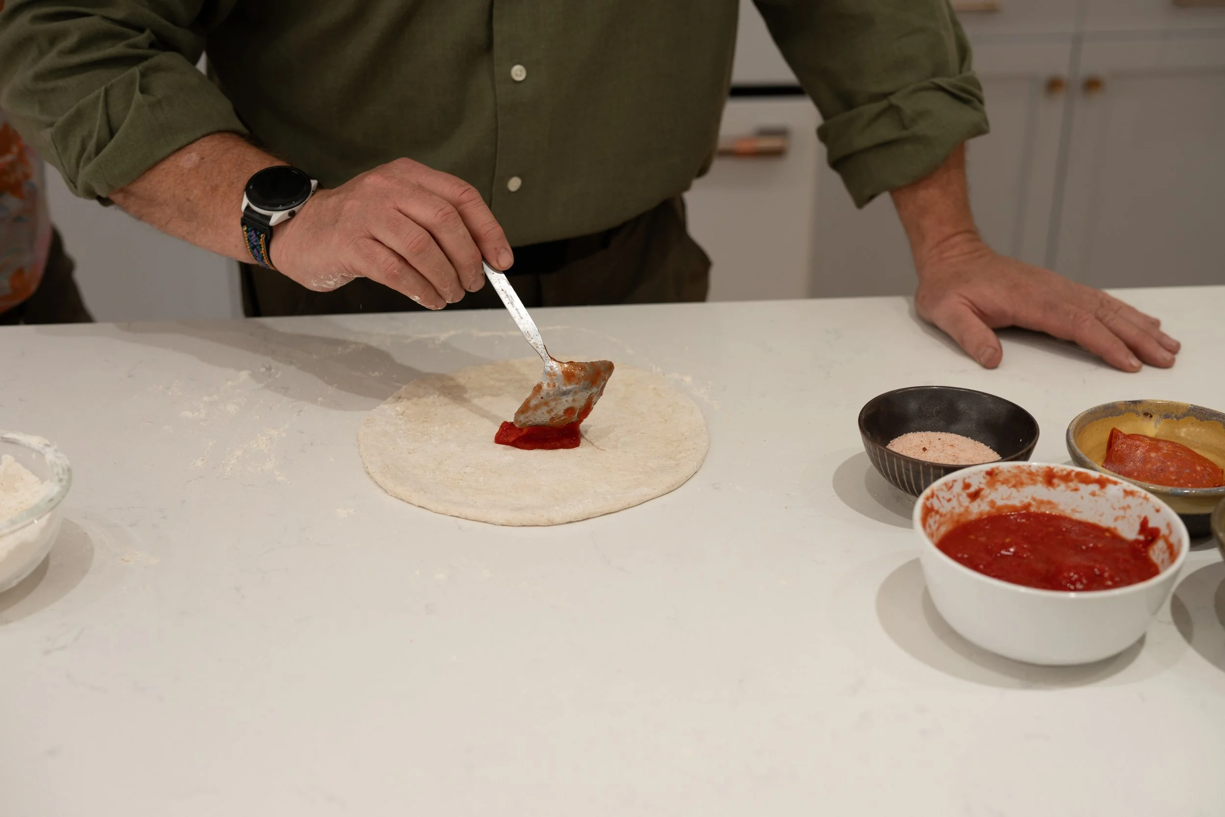 Person assembling a homemade pizza by spooning red sauce onto pizza dough with bowls of sauce and toppings nearby