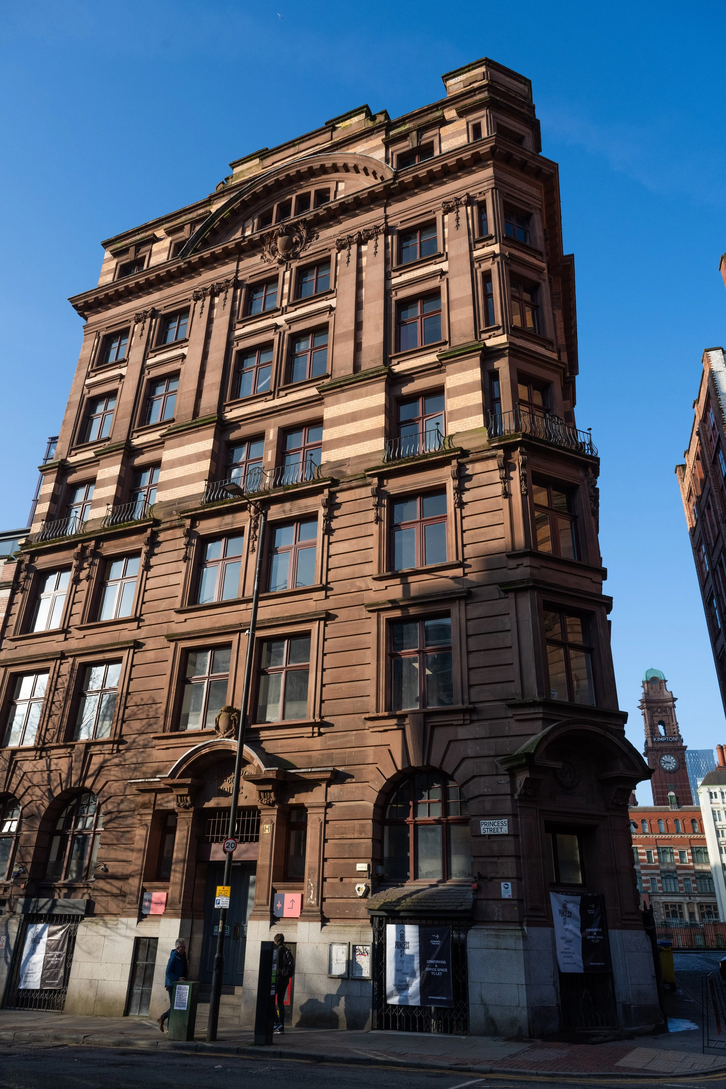 A tall historic building made of brown stone with multiple windows and balconies, with a clear blue sky in the background. There are street signs and people walking on the sidewalk in front of the building.