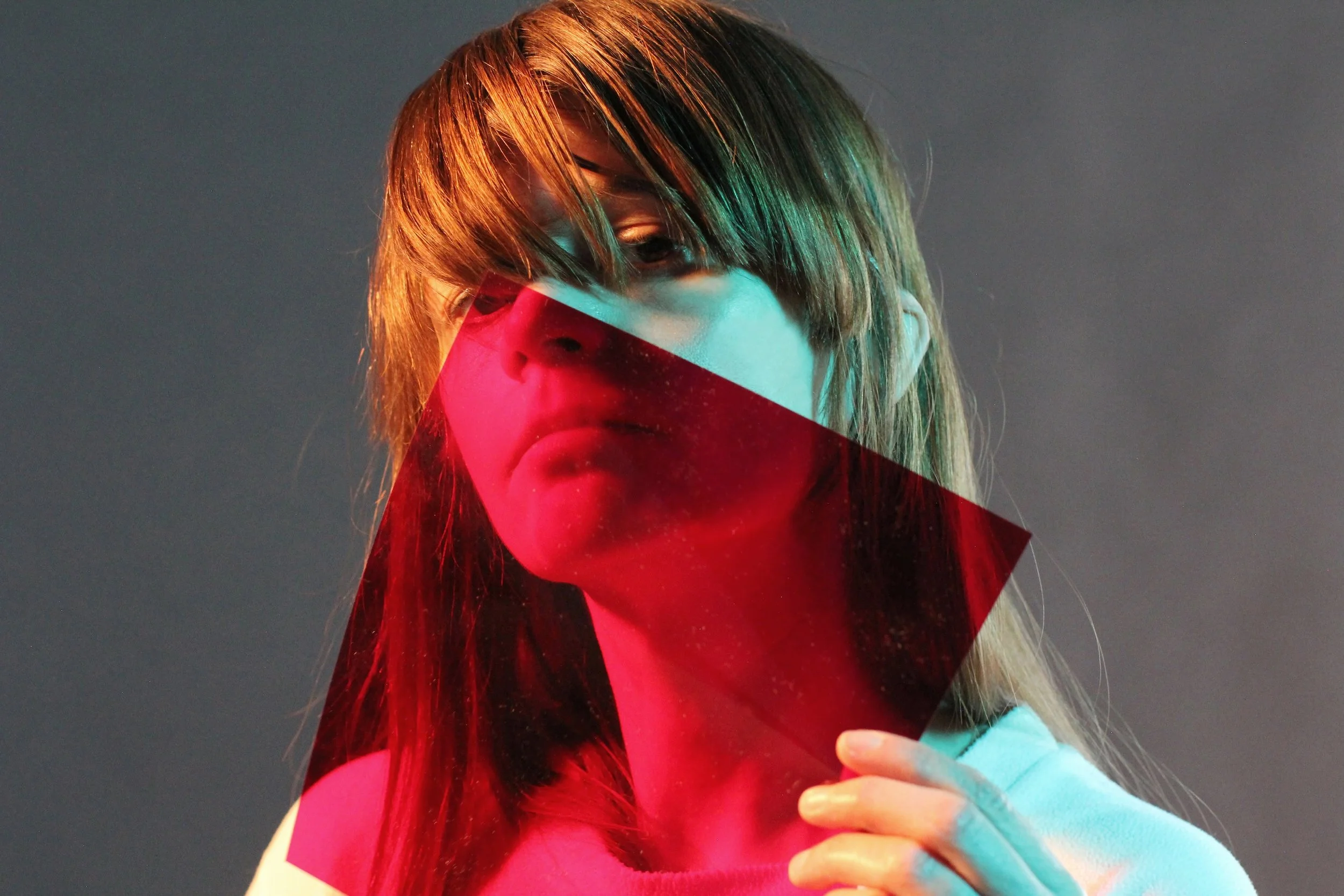 A woman with brown hair, wearing a mask with red and teal colors, holding a phone, against a dark background.