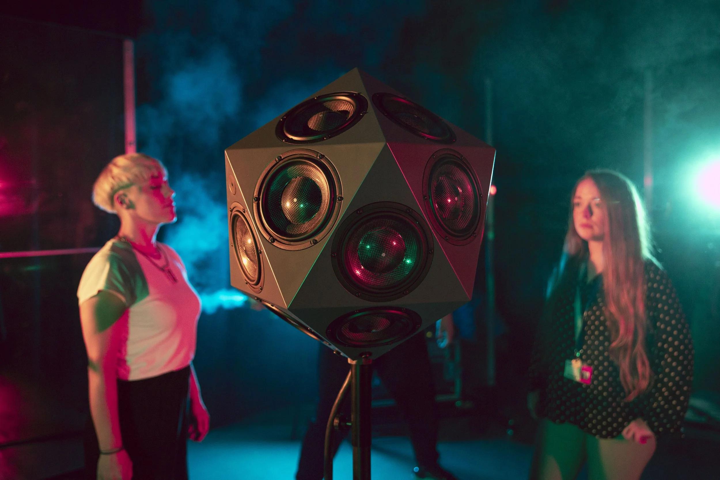 Two women standing on either side of a large, cube-shaped speaker with multiple circular drivers, in a dimly lit setting with colorful stage lighting and smoke, suggesting a music or dance event.