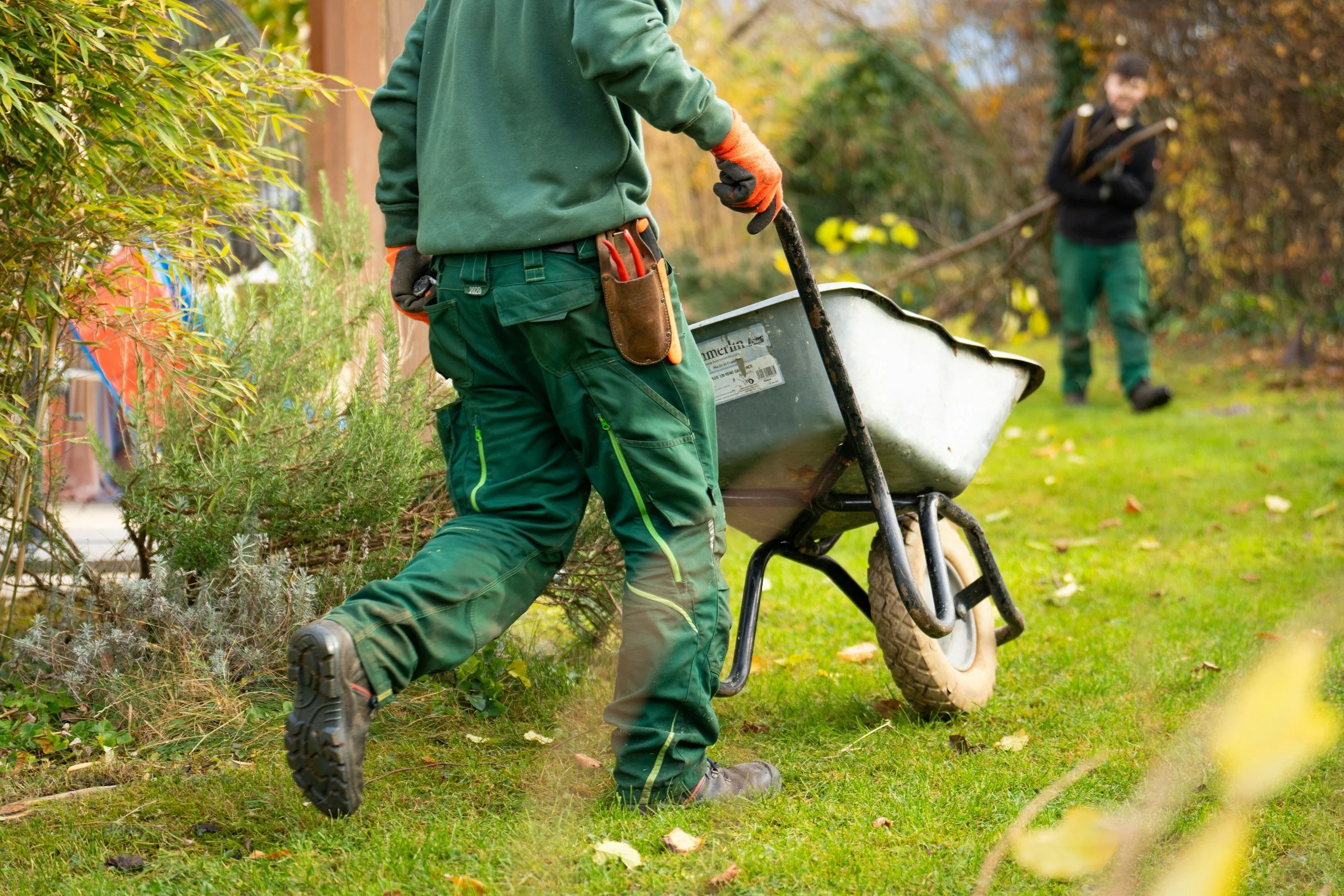 A person wearing green work pants and gloves is pushing a wheelbarrow in a garden. In the background, a young boy is holding a rake, standing among autumn-colored trees and plants.