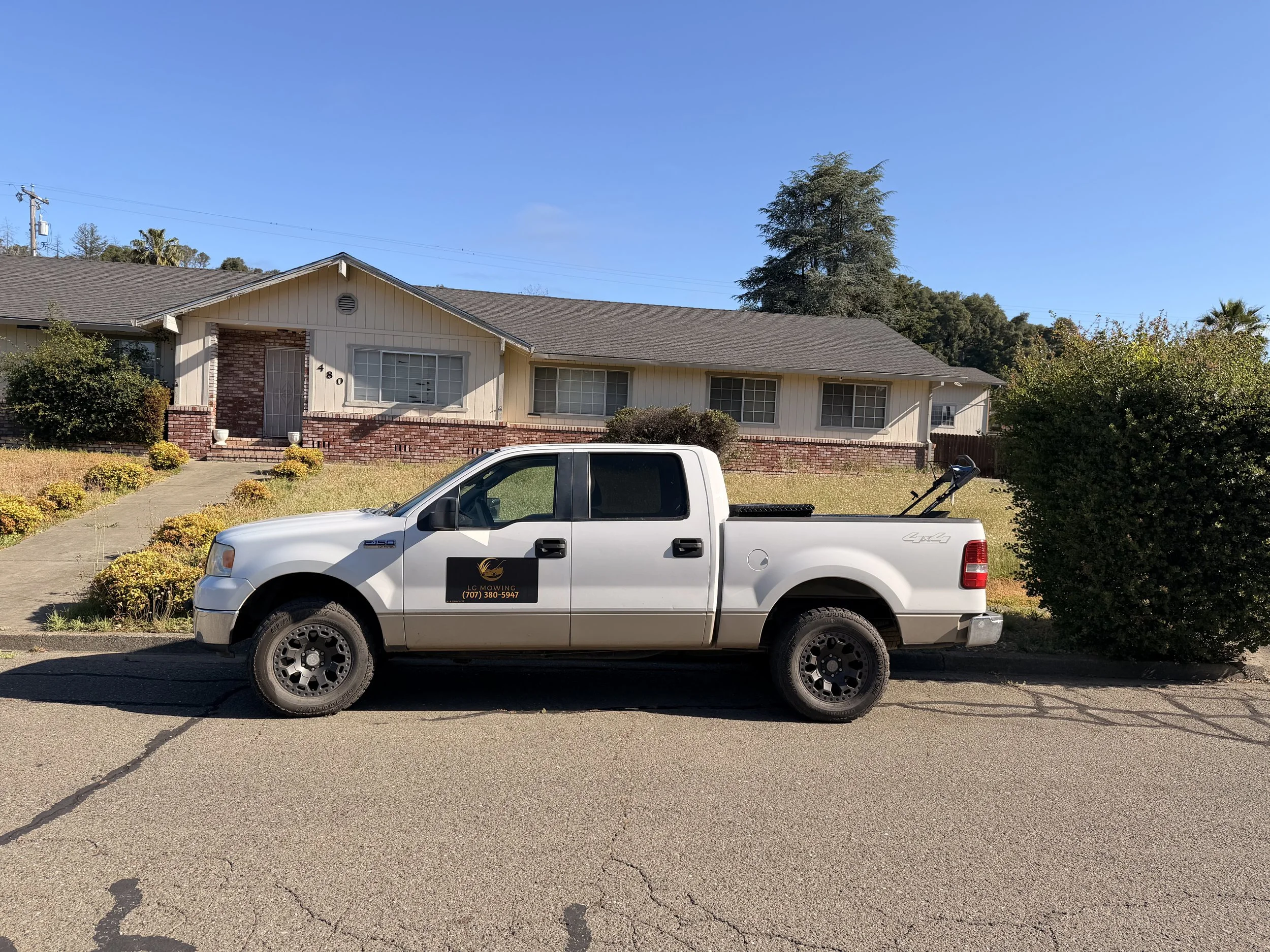 A white pickup truck parked on a driveway in front of a residential house. The house has a brick and cream-colored exterior with large windows. The yard has bushes and small plants, and trees are visible in the background under a clear blue sky.
