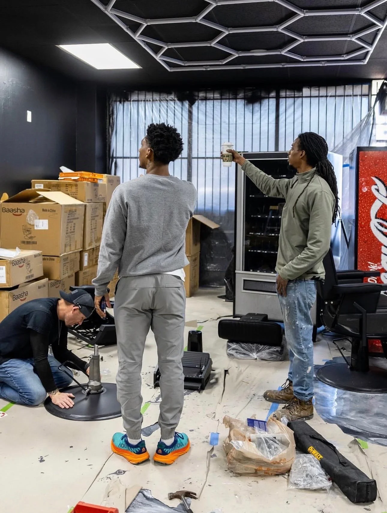 Three men working in a workshop or storage room, one of them wearing bright orange shoes, two of them holding tools, surrounded by boxes and equipment.