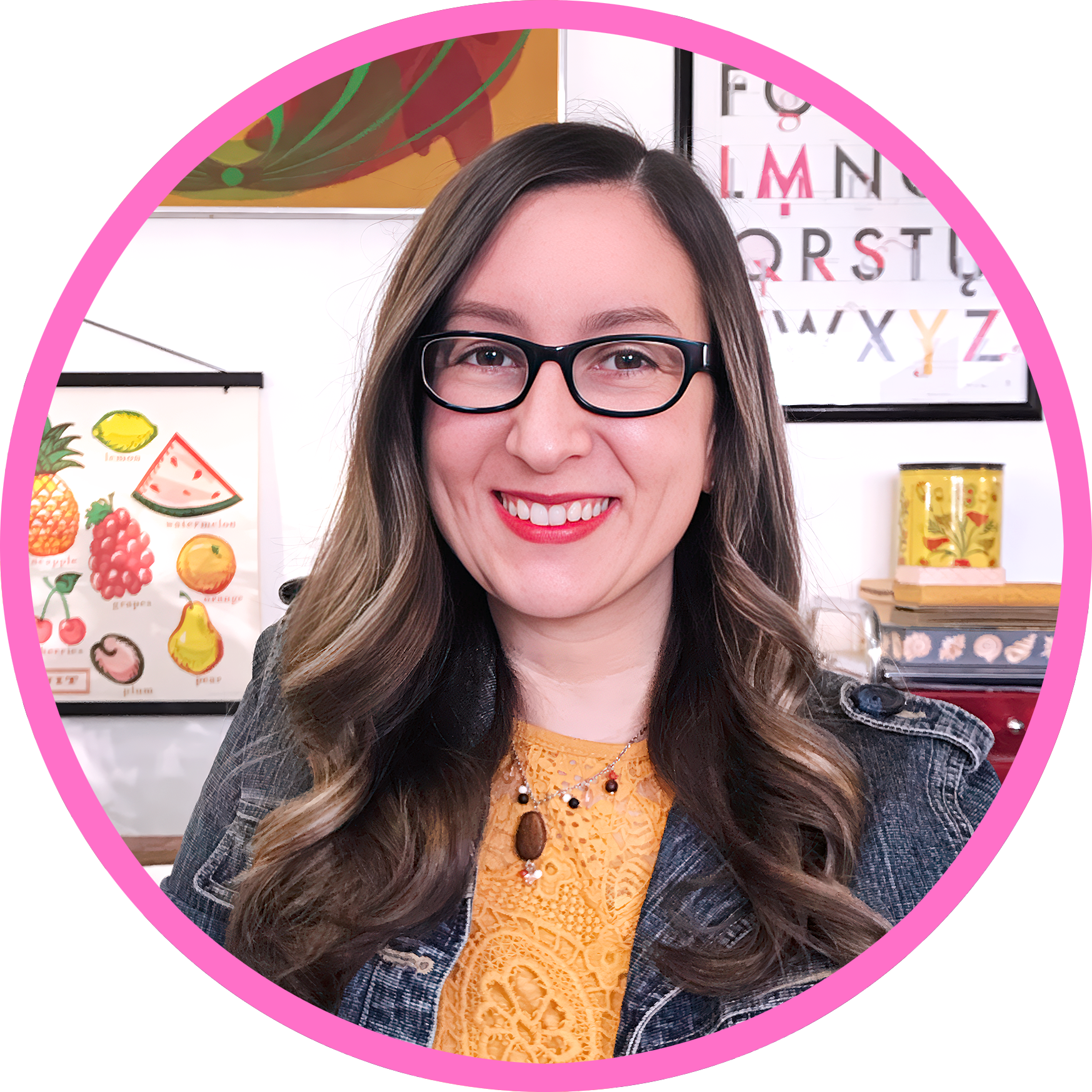 A woman with long wavy hair, glasses, and a mustard-colored top, smiling in an art studio setting with colorful artistic posters and decorations in the background.