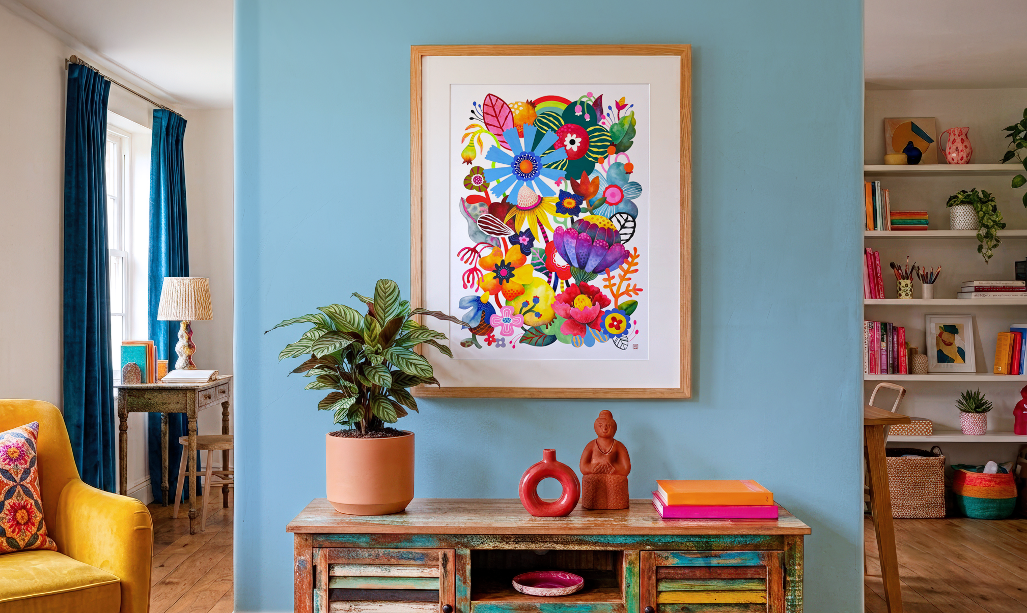 A colorful abstract botanical art print hangs on a light blue wall in a cozy, decorated living room. In the foreground, a wooden table displays three ceramic vases and sculptures, with a plant nearby. To the left, a yellow armchair with a patterned pillow, and to the right, a white bookshelf with books and decorative items. A window with blue curtains is also visible.