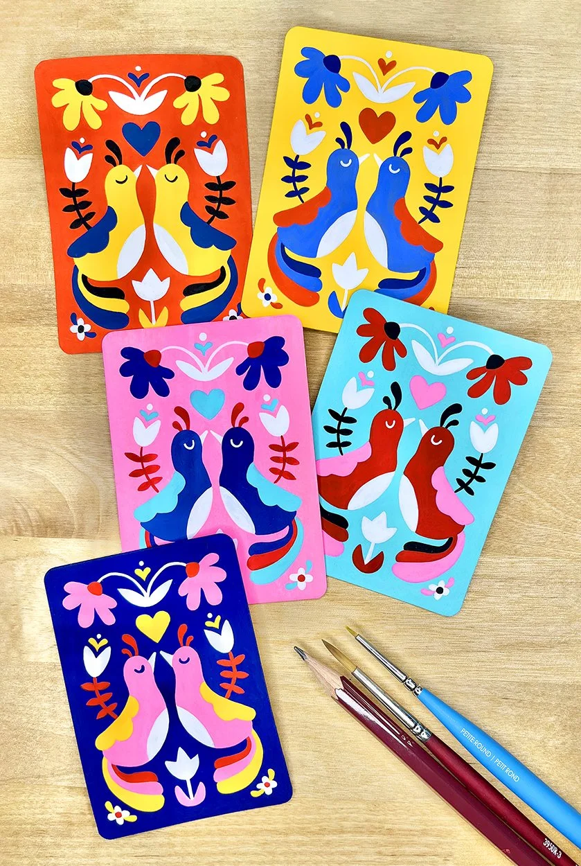 Five colorful illustrated cards on a wooden surface, each featuring two stylized rooster or bird figures surrounded by flowers, hearts, and decorative elements, along with two paintbrushes.