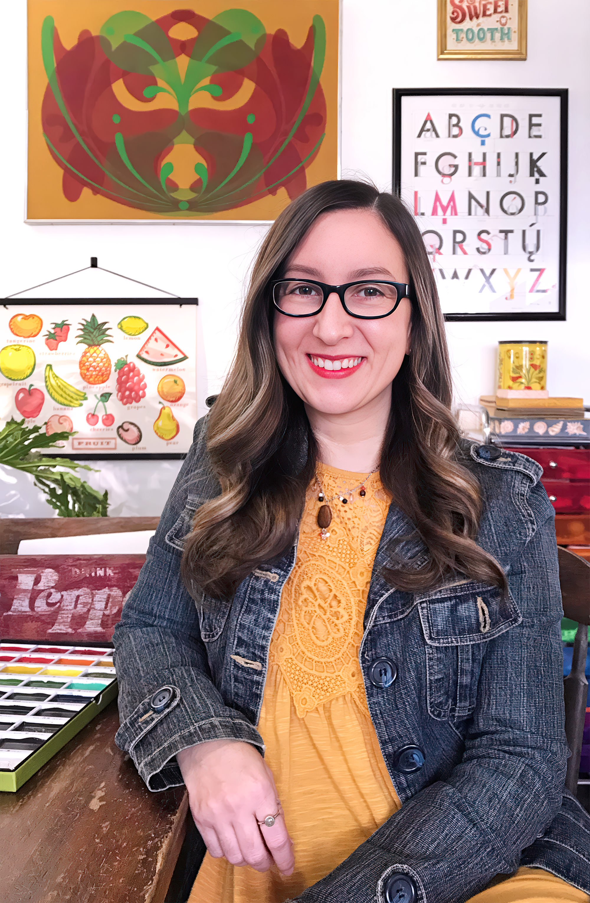 A woman with long wavy hair, glasses, and a mustard-colored top, smiling in an art studio setting with colorful artistic posters and decorations in the background.