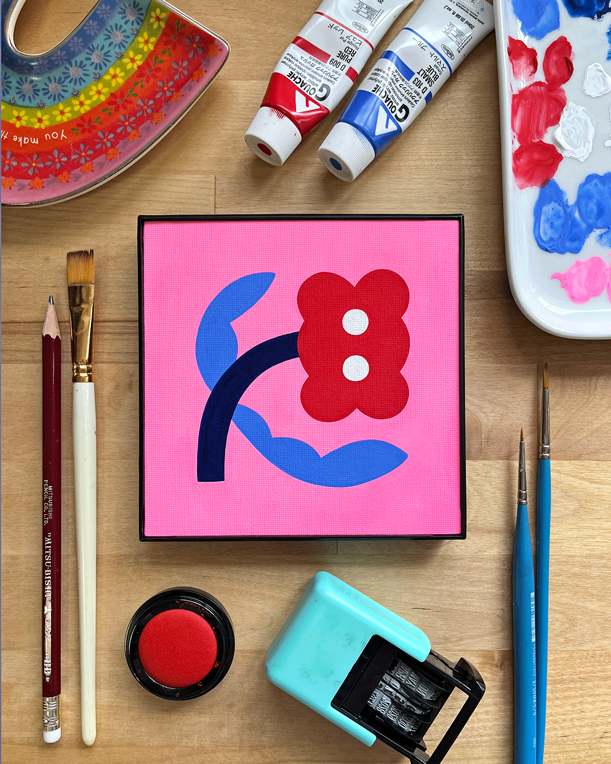 Colorful artwork on a digital screen depicting a stylized flower with red petals and white dots, set on a pink background, surrounded by painting supplies and tools.