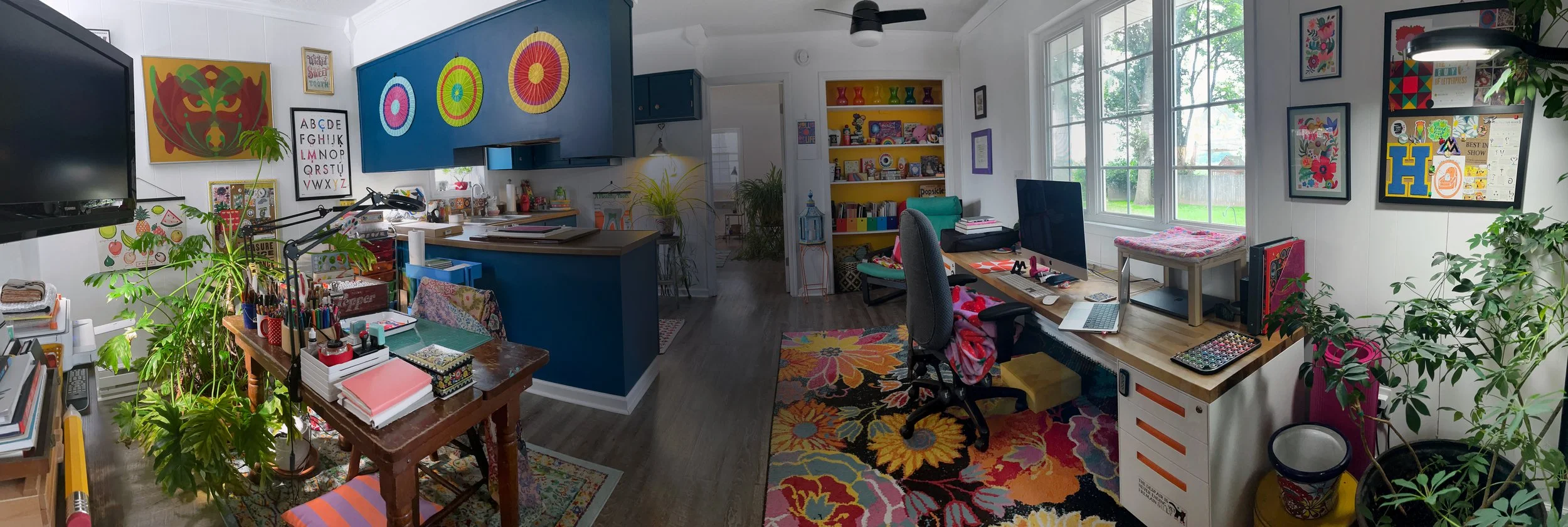 Colorful home office and craft room with large window, decorated with artwork, plants, and various supplies.