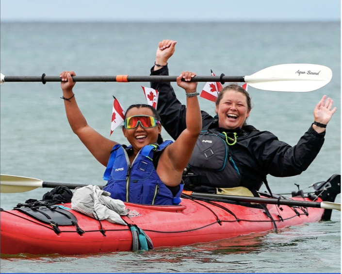 Two women in a red kayak on the water, smiling and celebrating, with one holding a paddle above her head. Both wear life jackets and Canadian flags on their hats, with a cloudy sky in the background.