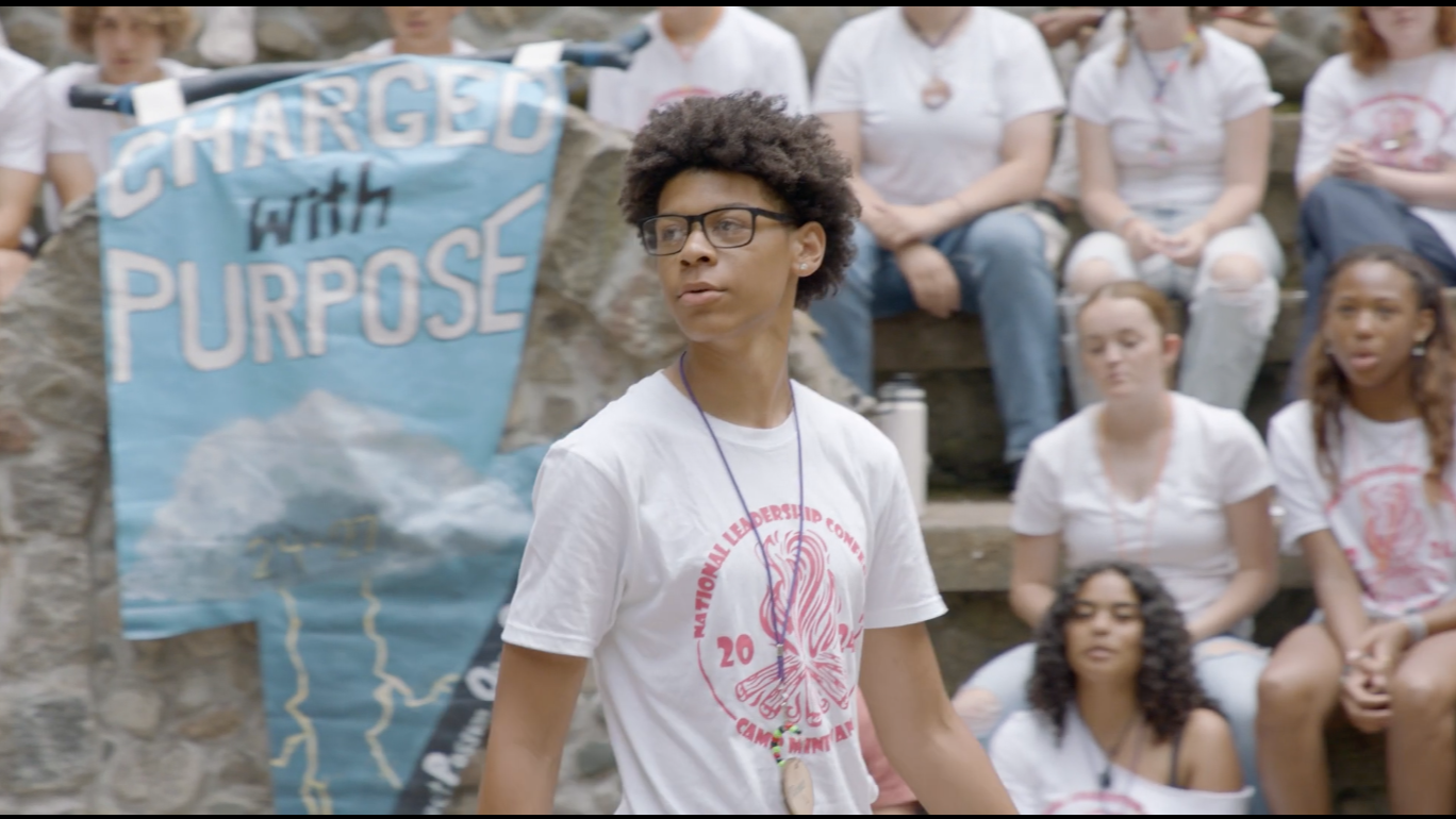 A young man with short curly hair, glasses, and a white t-shirt with a red logo, stands among seated teens at an outdoor gathering. Behind her, there is a blue banner with the words 'CHARGED with PURPOSE' and a group of women sitting on stone steps.