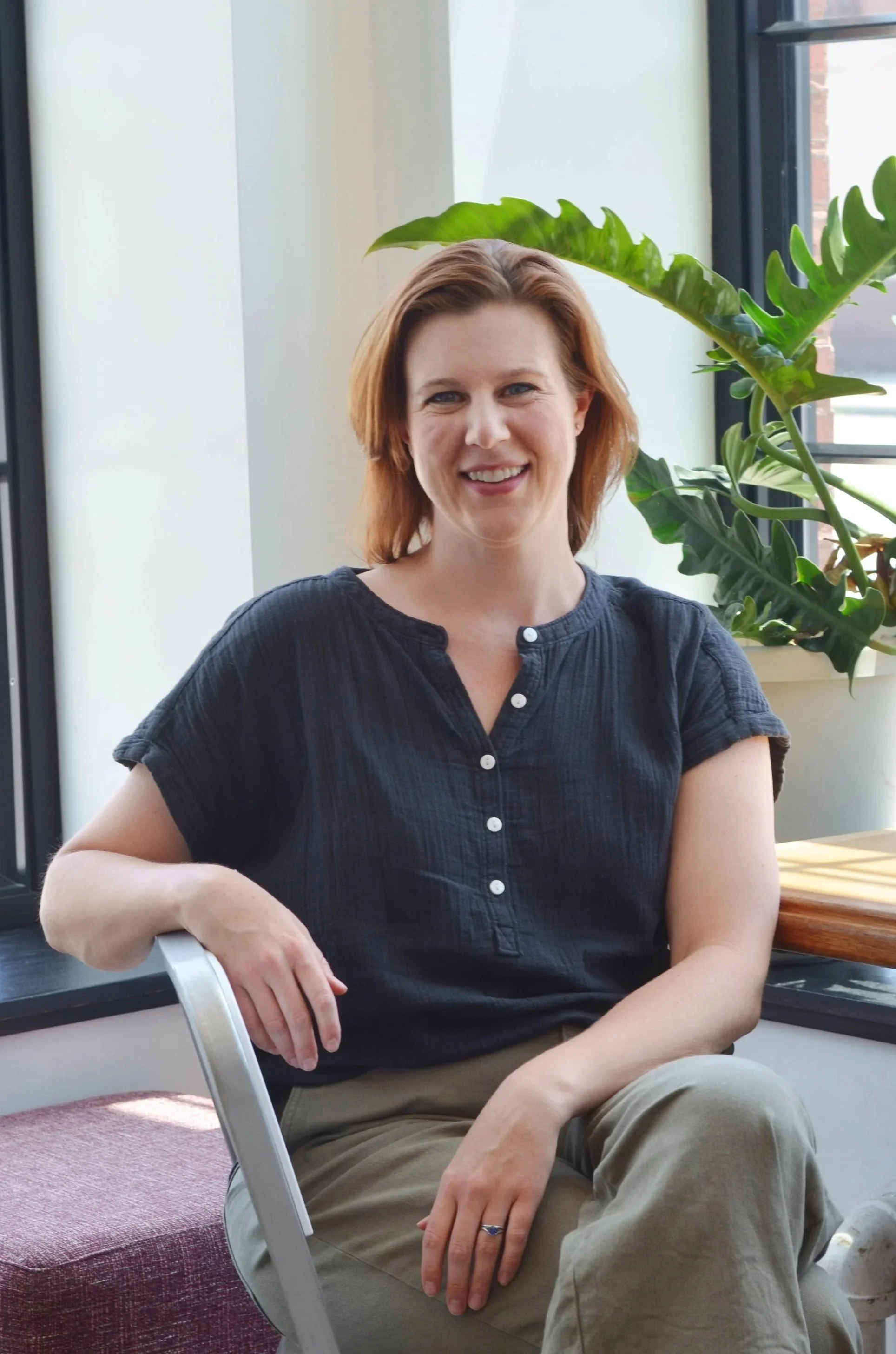 A woman with short red hair smiling, sitting near a large window with green plants behind her, wearing a dark short-sleeved shirt and beige pants.