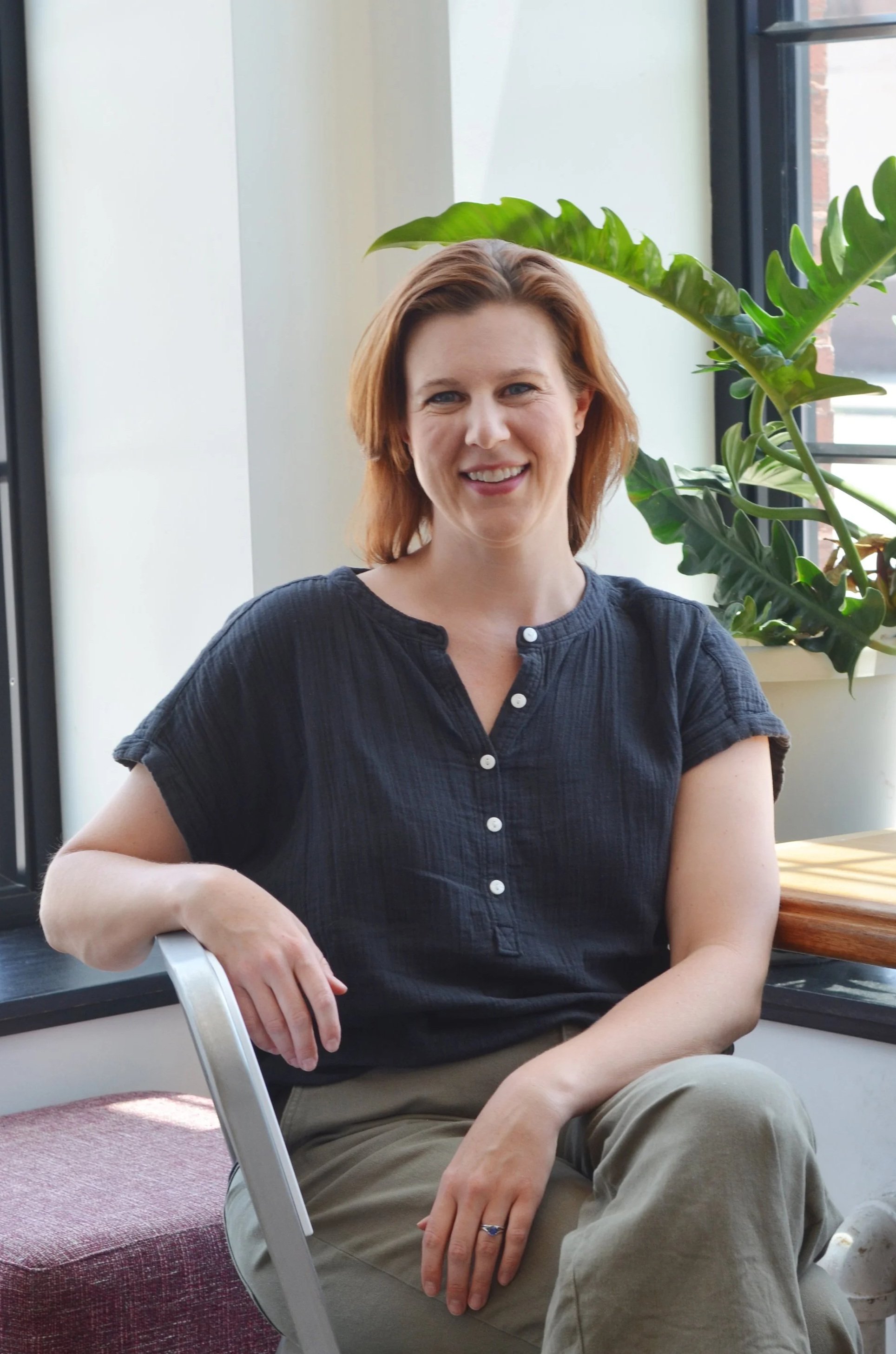 A woman with short red hair sitting indoors near large windows and green plants, smiling at the camera.