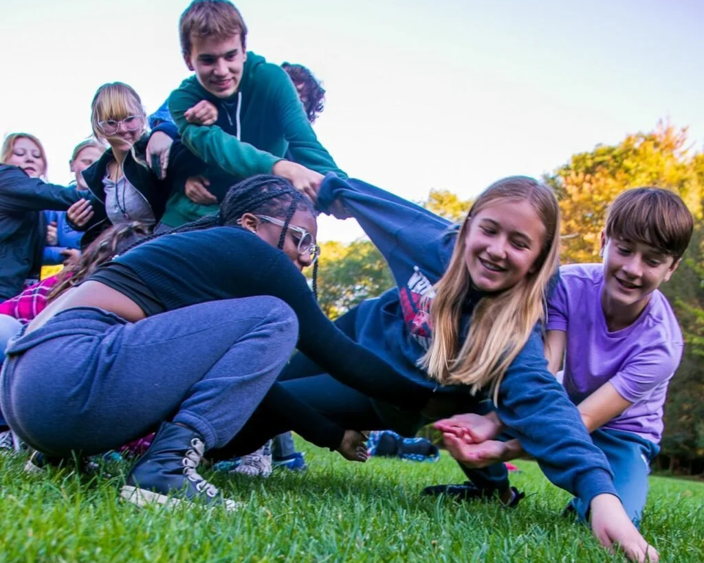 A group of kids participating in a team building exercise outdoors on a grassy field during daytime with trees and a clear sky in the background.