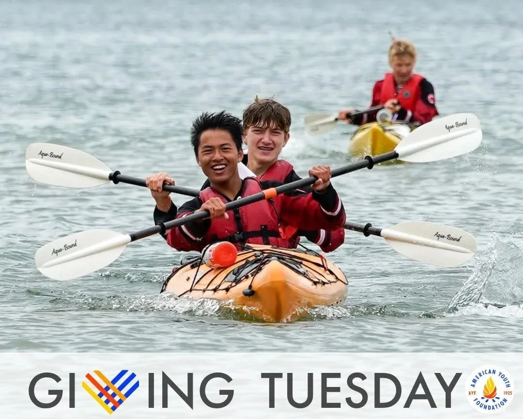 Three young people kayaking on a lake with smiles, wearing red life jackets, in yellow and orange kayaks, paddling with white oars.
