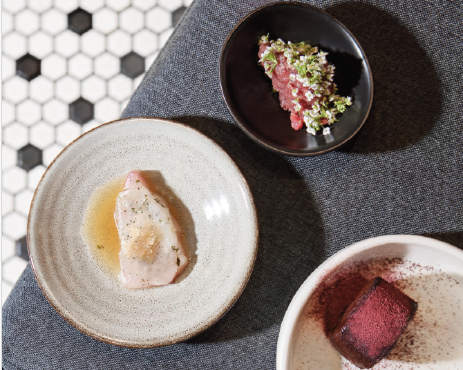 Three small dishes with gourmet food, including a piece of white fish in sauce, a pink meat or tartare with herbs, and a chocolate dessert with red powder, on a dark fabric surface.