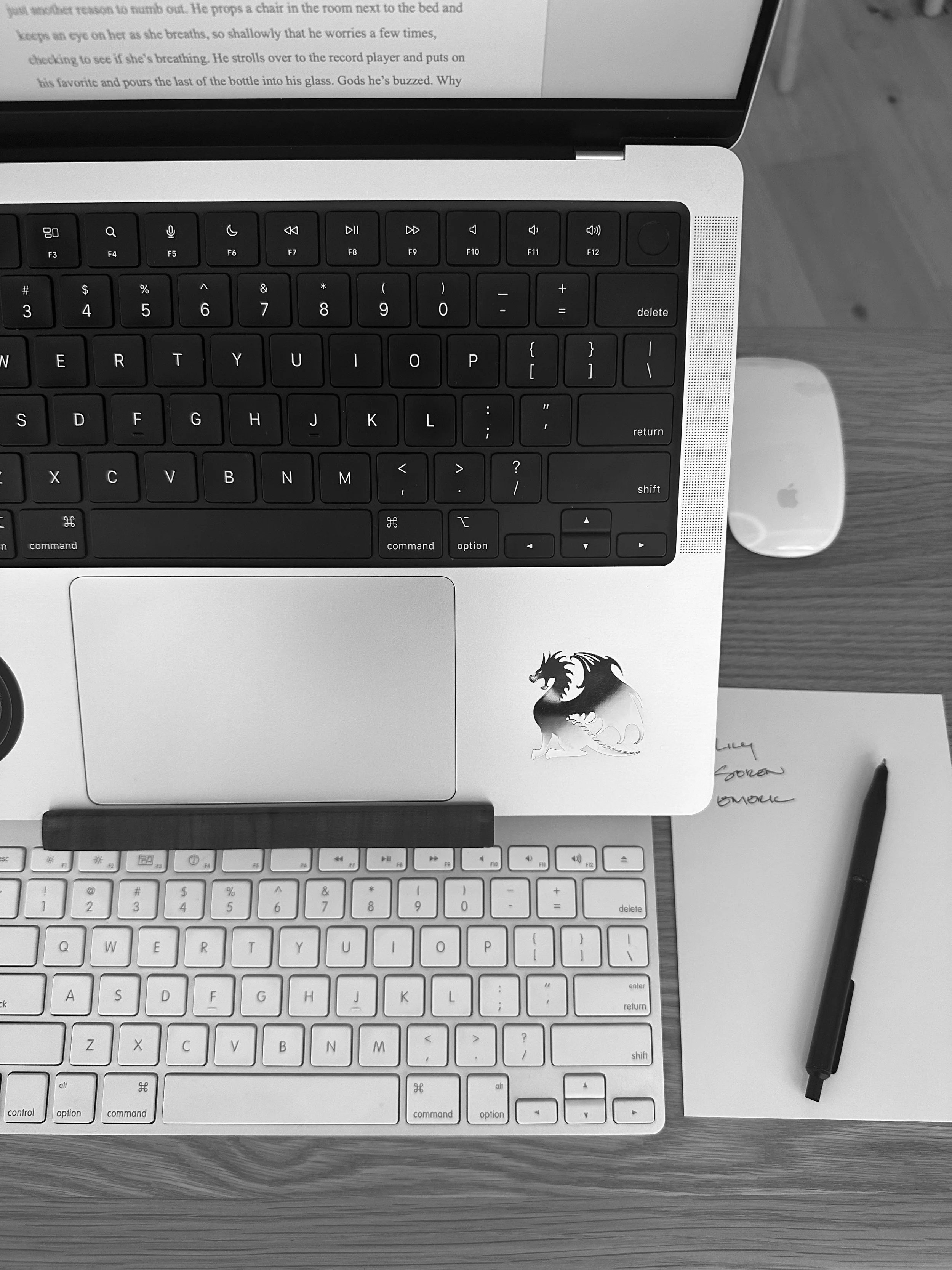 A flat lay image of various electronic devices, including a laptop, a white keyboard, and a wireless mouse, placed on a wooden desk with a piece of paper and a pen nearby.