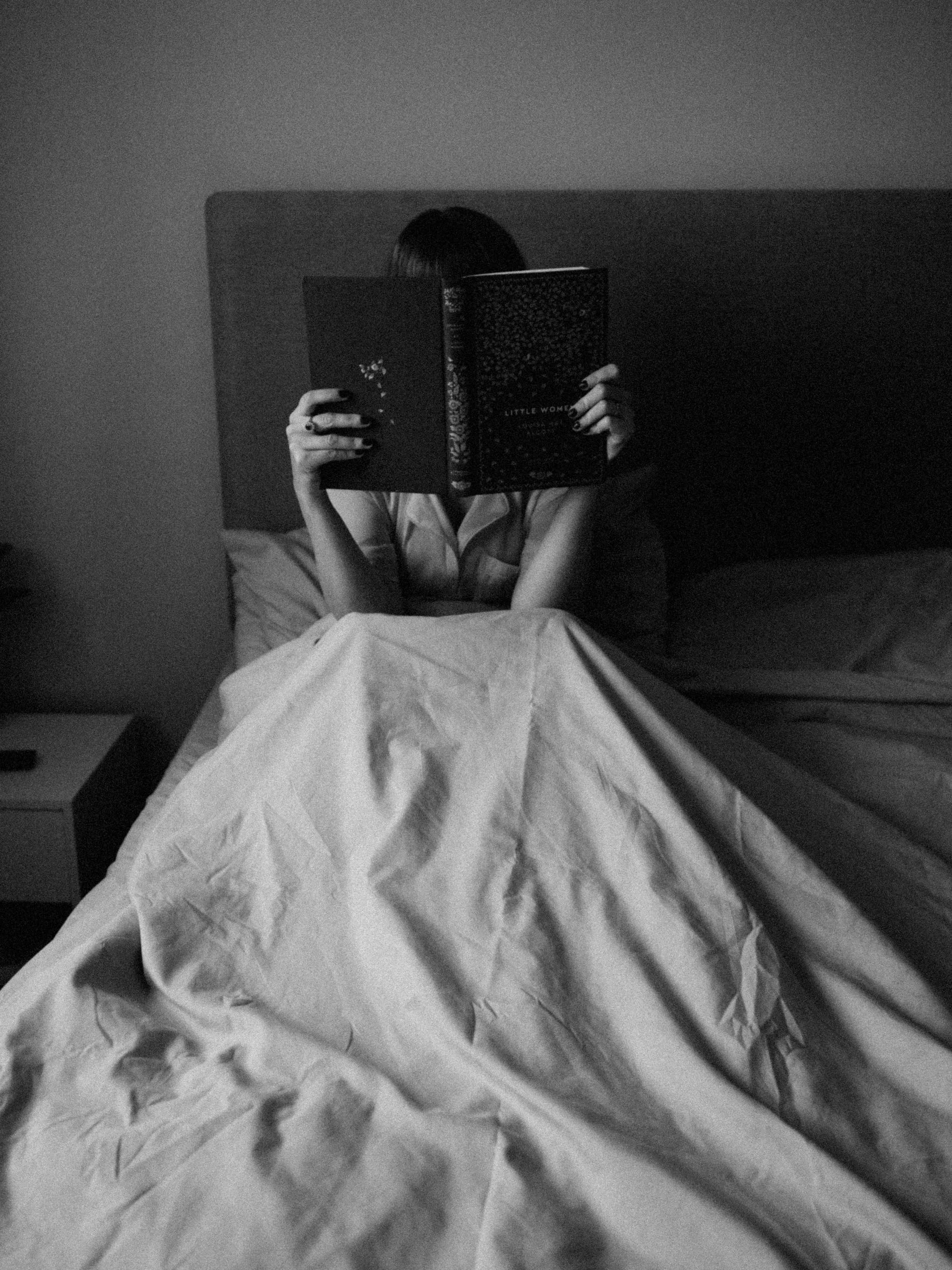 A person sitting on a bed, reading a book titled 'Little Women', in a dimly lit room with a plain wall in the background.