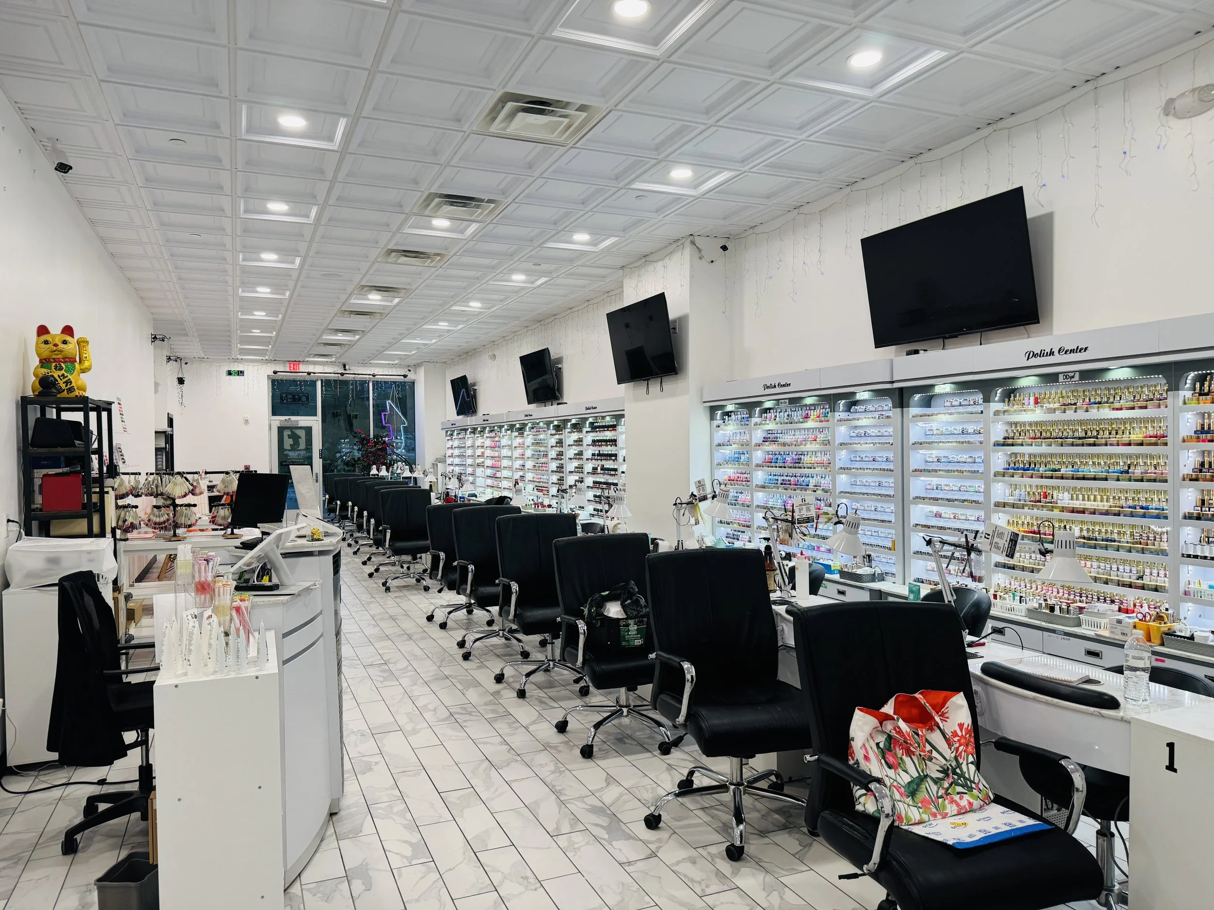 Interior of a nail salon with white tiled floor, white walls, and rows of black salon chairs. Multiple large flat-screen TVs are mounted on the wall. Shelves stocked with nail polish fill the space. There are decorative string lights on the wall and a Maneki-neko with a raised paw on a black shelf to the left. Several workstations have manicure supplies, and a tote bag with a floral pattern is on one of the chairs.