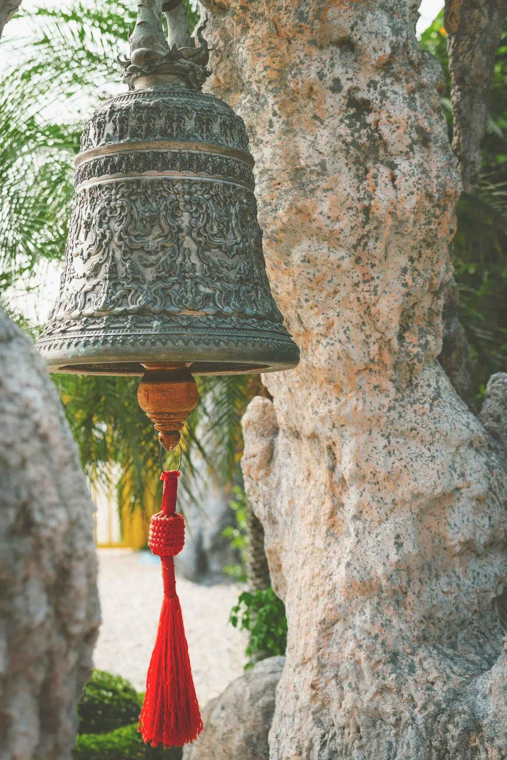 A decorative bell with intricate carvings hangs from a wooden support on a tree with textured bark, against a backdrop of green leaves and a bright outdoor setting.