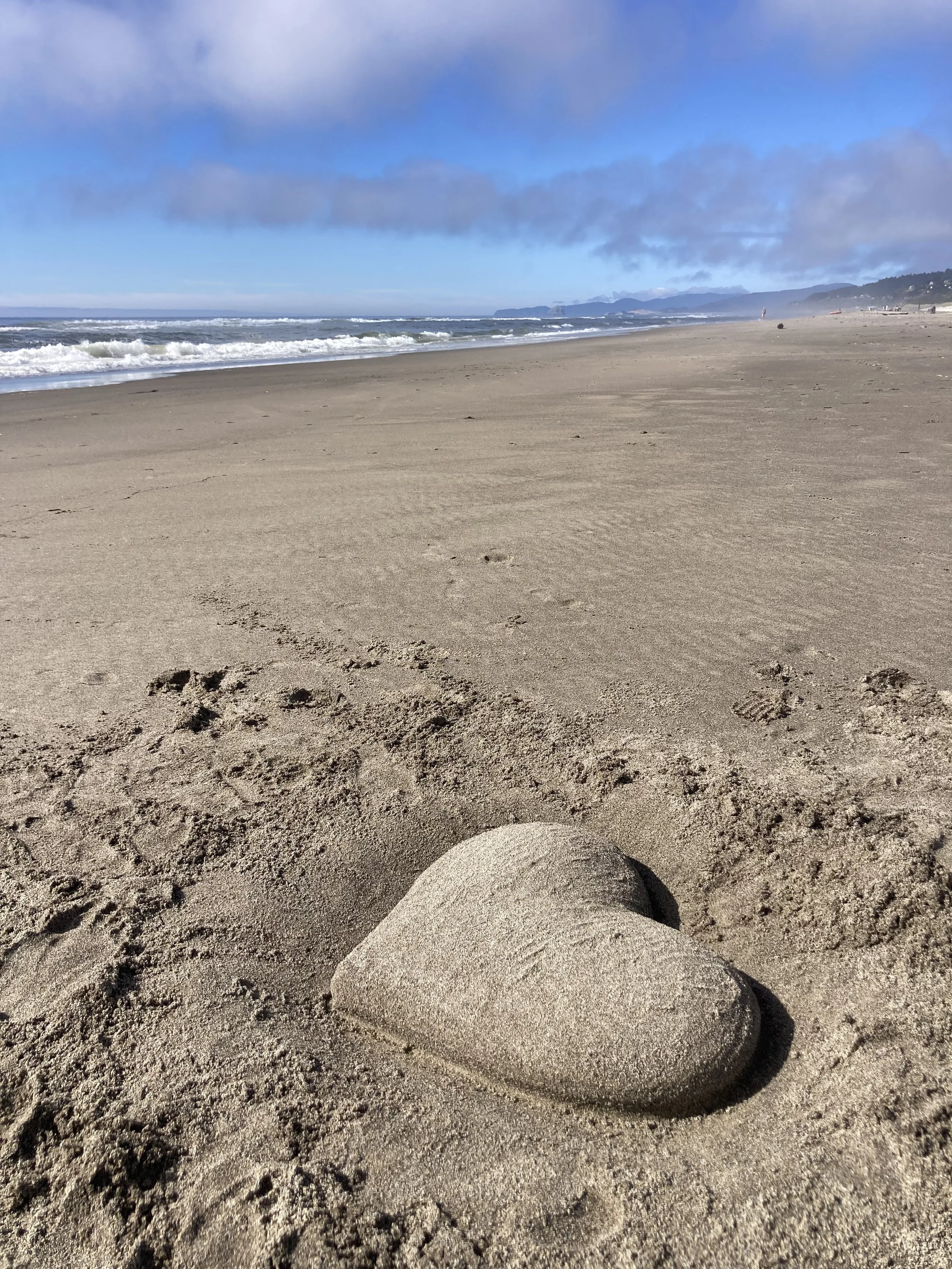 A sandy beach with a heart shaped out of sand in the foreground, ocean waves, a person in the distance, and a cloudy sky.