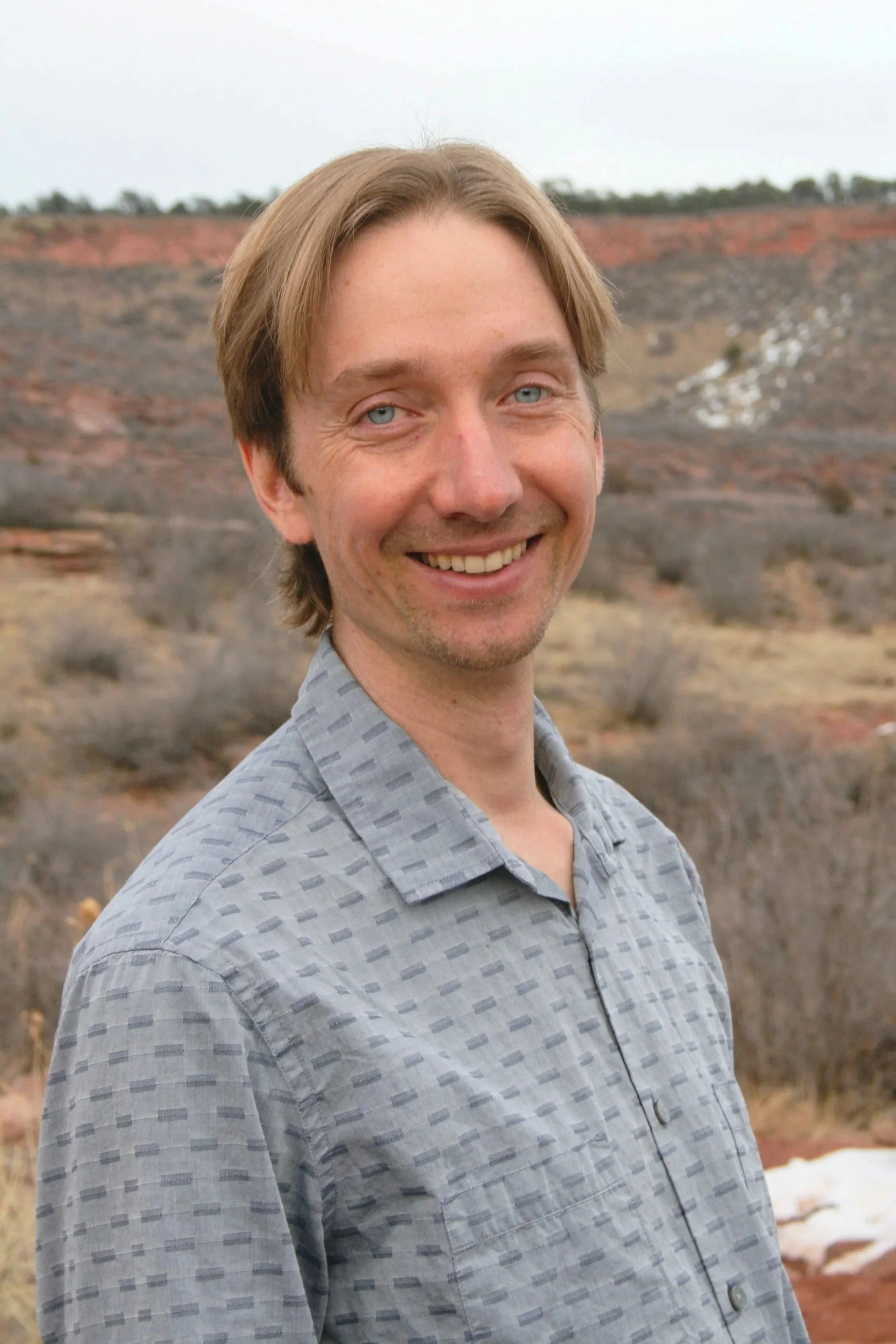 Sean Arnold smiling outdoors in a mountainous area with sparse vegetation and rocks in the background.