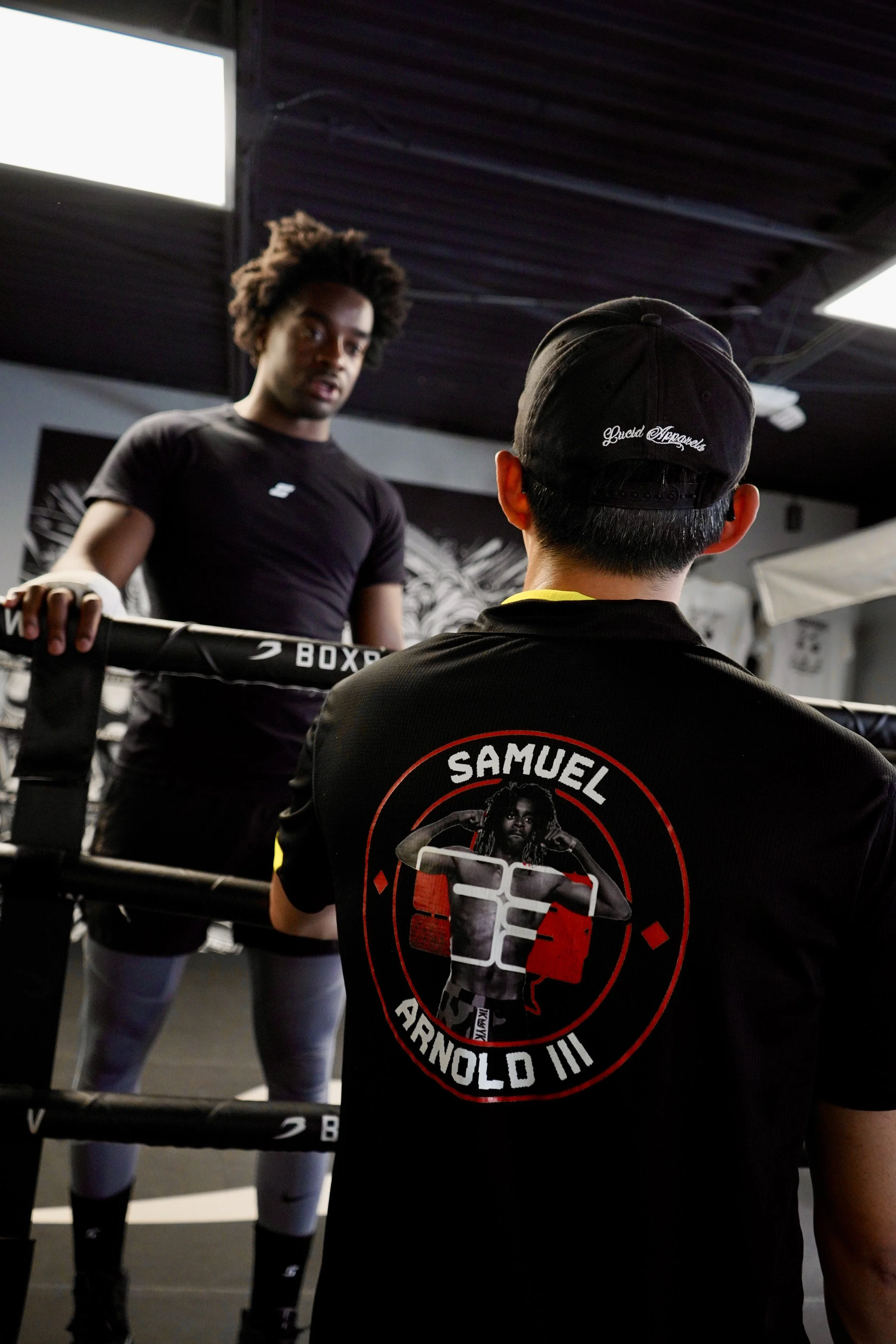 A young man with curly hair coaching a boxer in a gym. The boxer is wearing a black shirt with a graphic print on the back. The gym has a black ceiling and boxing ring ropes.