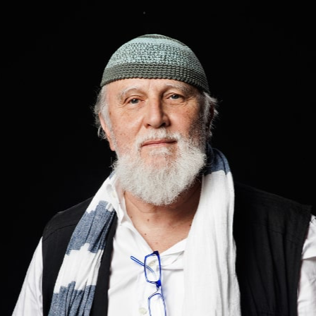 Portrait of an elderly man with a white beard, wearing a gray knit cap, glasses hanging from his shirt, a white scarf, and a black vest against a black background.