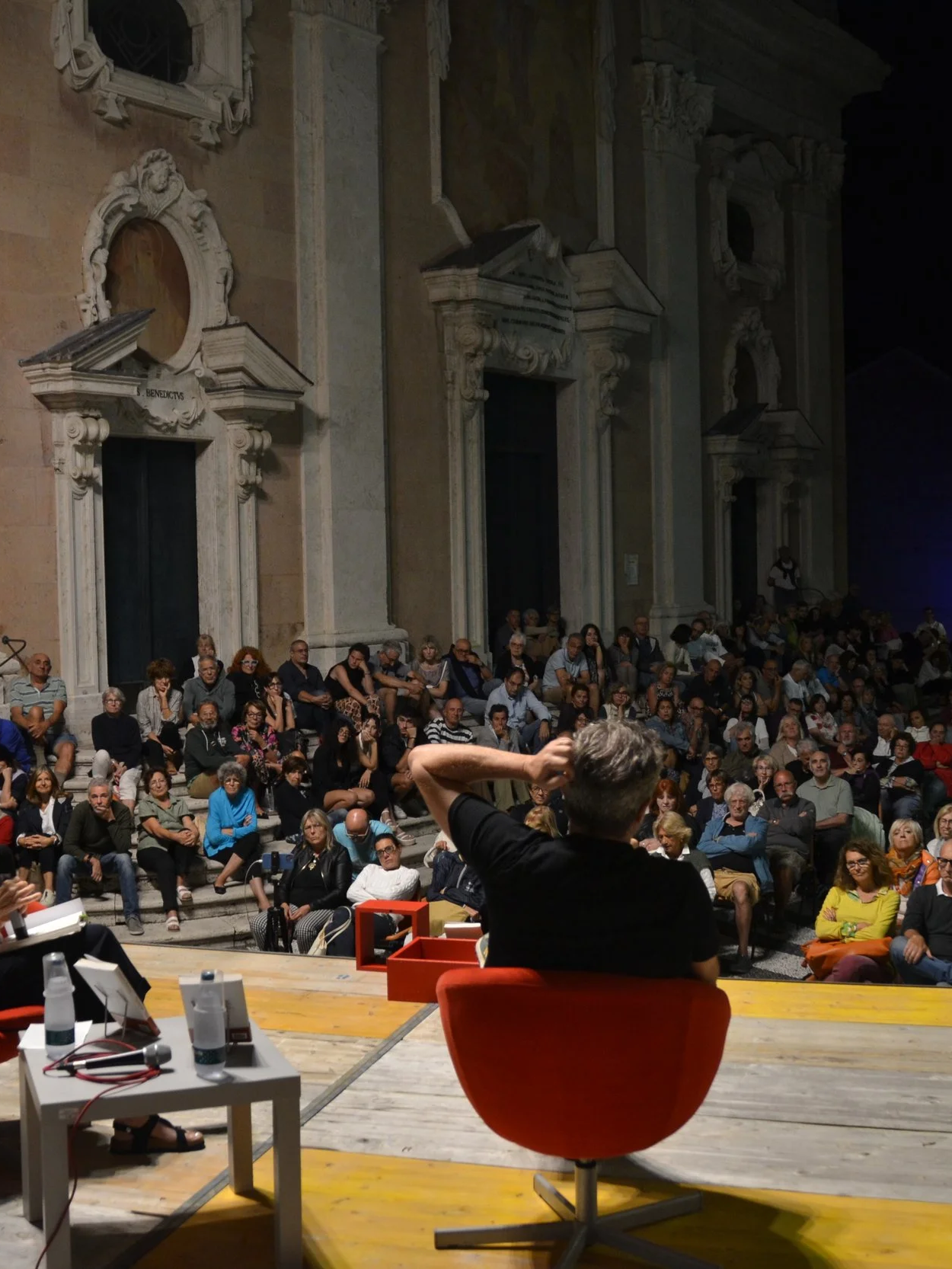 A large audience seated on steps and chairs inside an ornate, historic building listening to a speaker. The image is taken from the stage where a person with gray hair is seated, with audio-visual equipment and a table with water bottles in view.