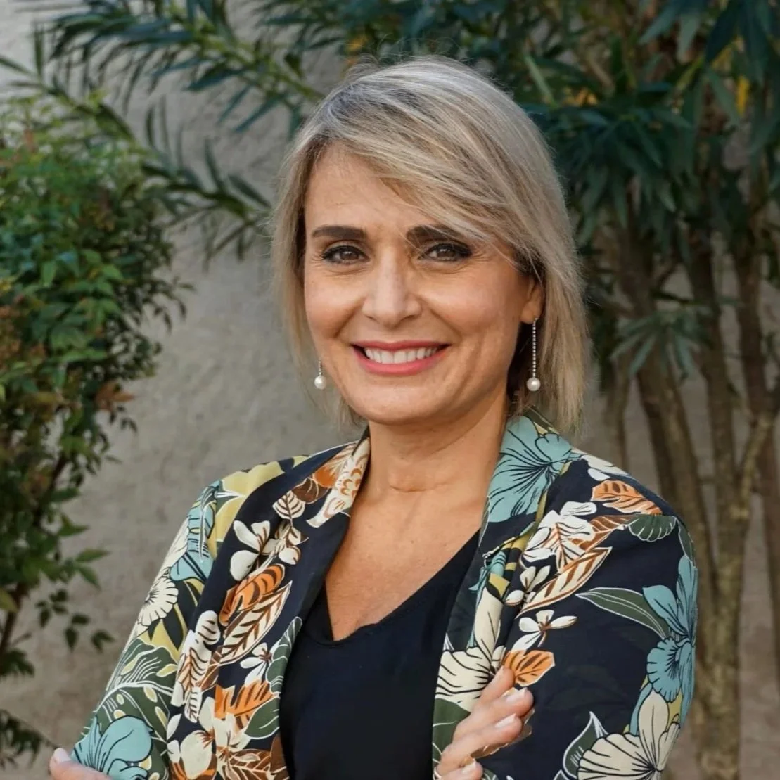 A smiling woman with blonde hair and pearl earrings standing outdoors in front of green plants, wearing a floral patterned blazer over a black top.