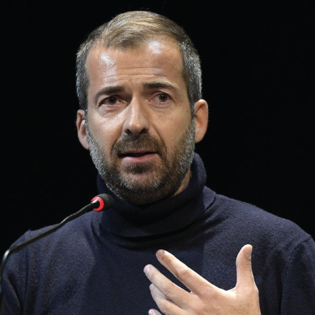 A man with a beard and short hair, speaking into a microphone and wearing a dark turtleneck sweater, gesturing with his hand.