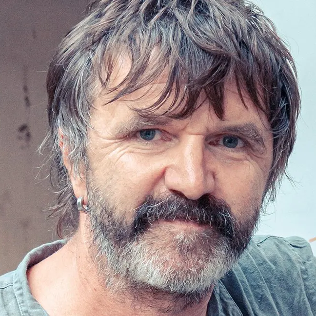 Close-up image of a middle-aged man with grayish-brown hair and a full beard, wearing earrings and a casual shirt.