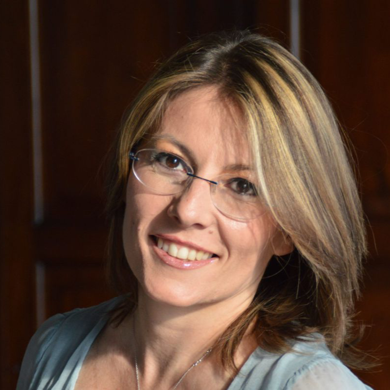 A woman with shoulder-length light brown hair with blonde highlights, wearing glasses with thin frames, smiling at the camera, against a dark wooden background.