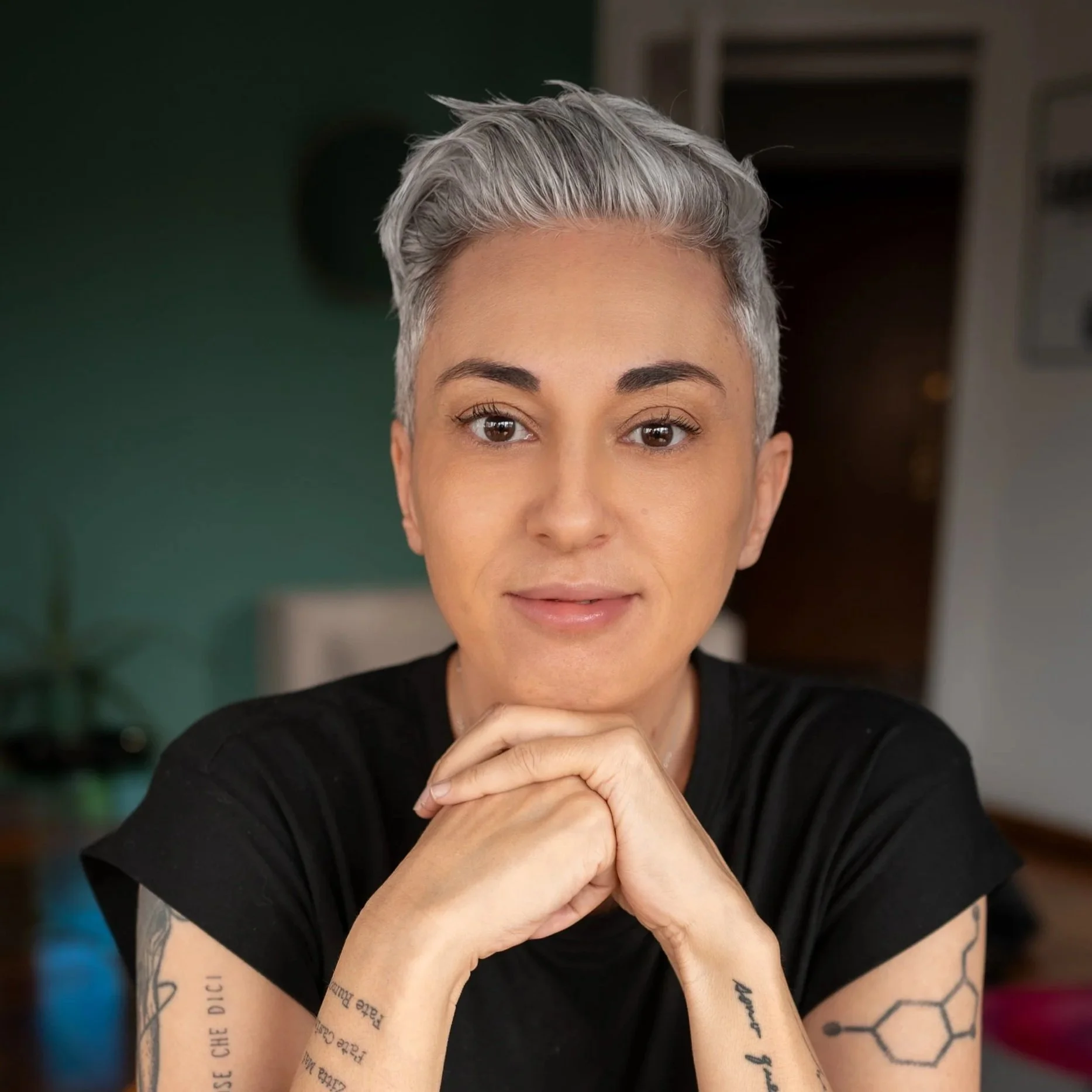 Close-up of a woman with short, silver-gray hair, expressive brown eyes, and tattoos on her arms, wearing a black top, sitting indoors with a green wall and furniture in the background.