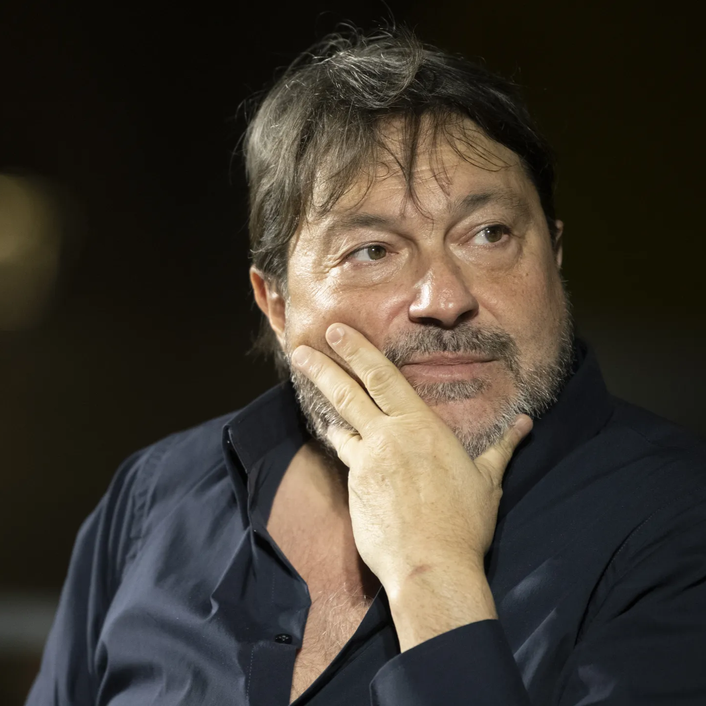 A man with gray hair and beard, wearing a dark shirt, resting his chin on his hand, looking thoughtful.