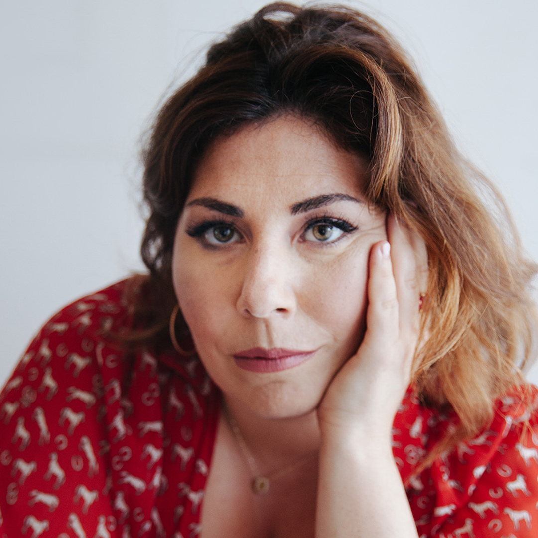 A woman with shoulder-length wavy red hair, wearing a red top with white patterns, resting her face on her hand and looking thoughtful.