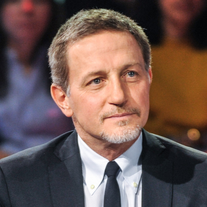 A man with short light brown hair, a trimmed goatee, wearing a dark suit, white shirt, and dark tie, sitting in a studio or courtroom setting, with blurred audience or panel members in the background.
