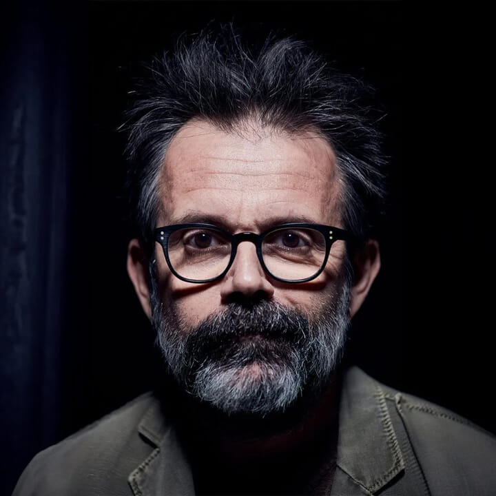A middle-aged man with salt-and-pepper hair and beard, wearing black-rimmed glasses, facing the camera against a dark background.