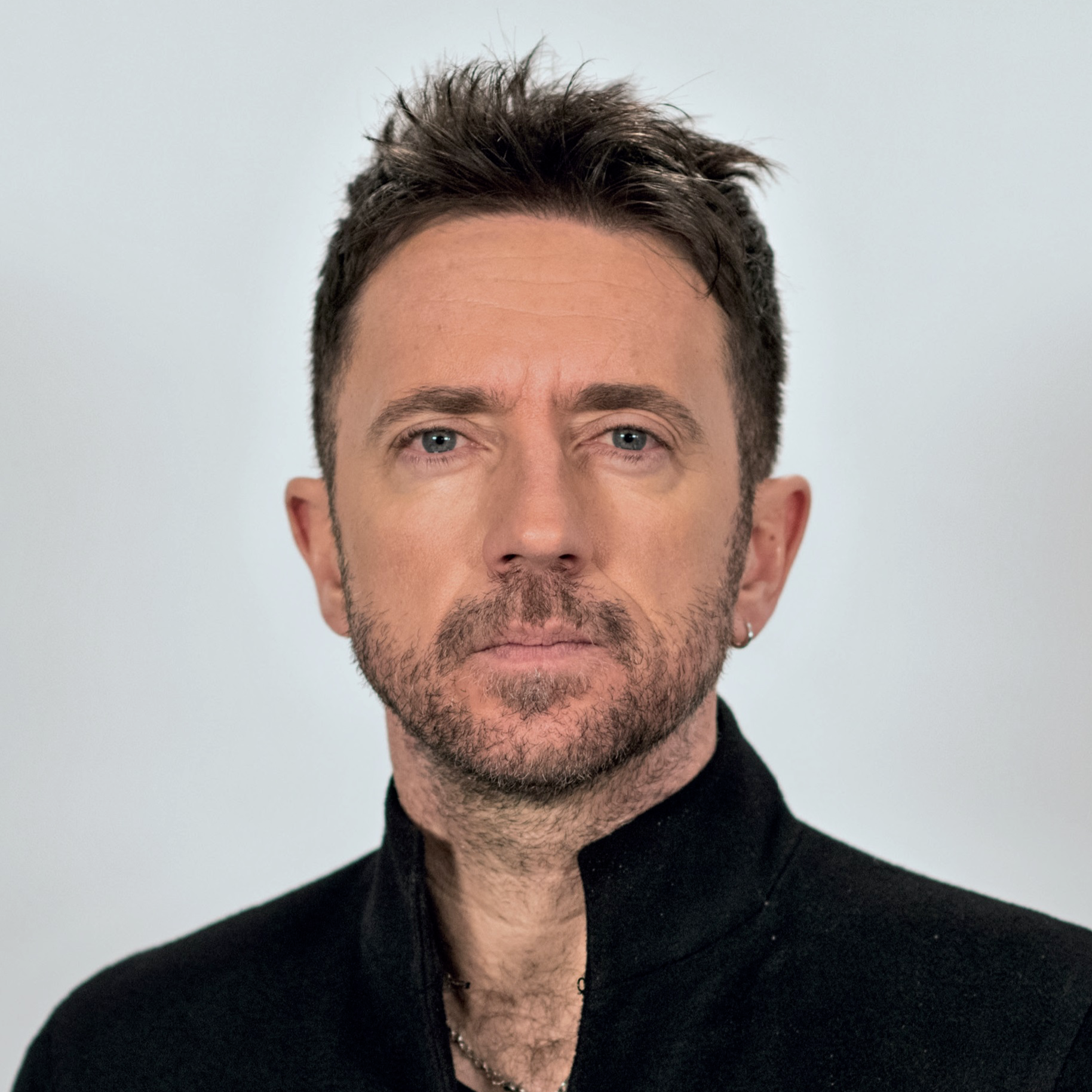 Close-up portrait of a man with blue eyes, short dark brown hair, facial hair, and a small earring in his left ear, wearing a black shirt and chain necklace against a plain light background.